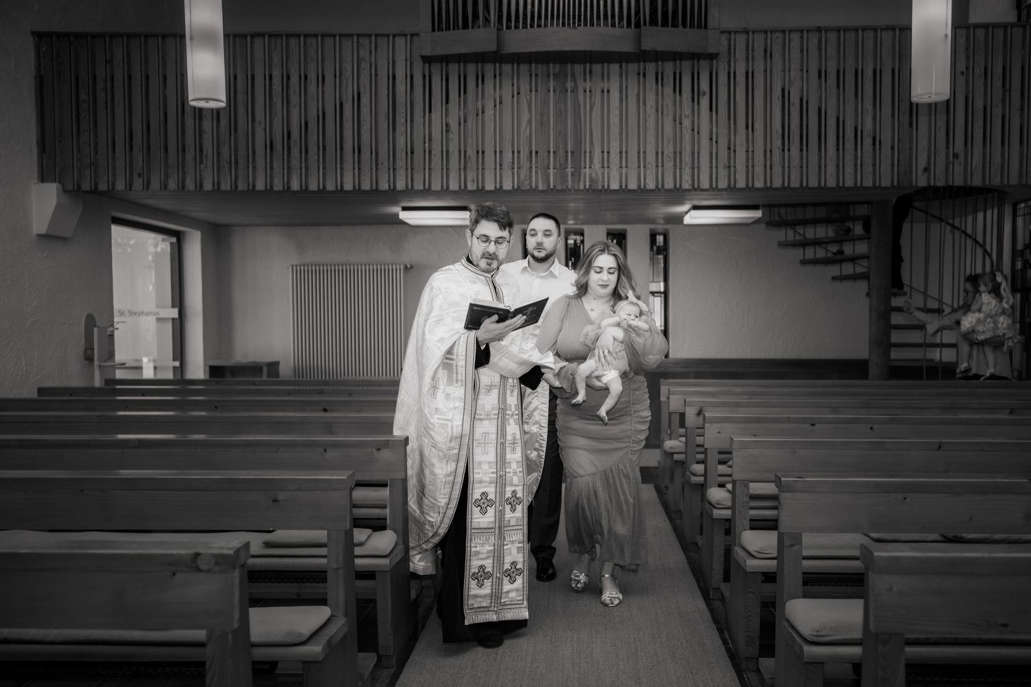 Schwarz-Weiß-Aufnahme der Familie mit Lara auf dem Arm und dem Priester im Gang zwischen den Kirchenbänken.