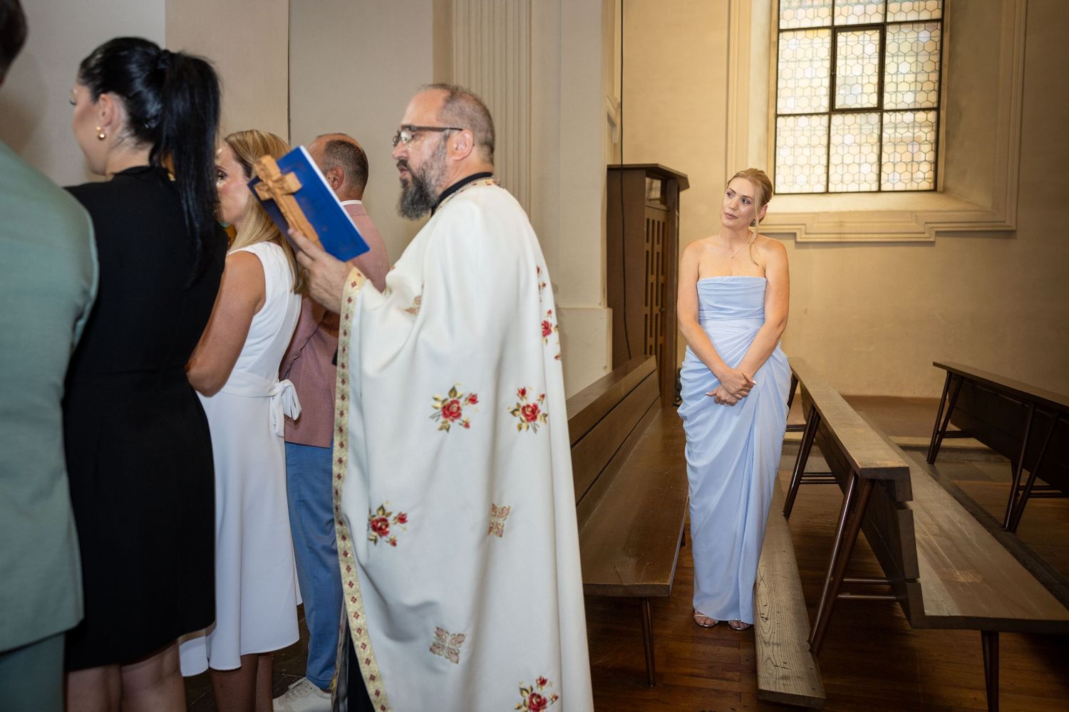 Eine Frau im hellblauen Kleid steht zwischen den Kirchenbänken; der Priester hält ein blaues Buch und ein Holzkreuz.