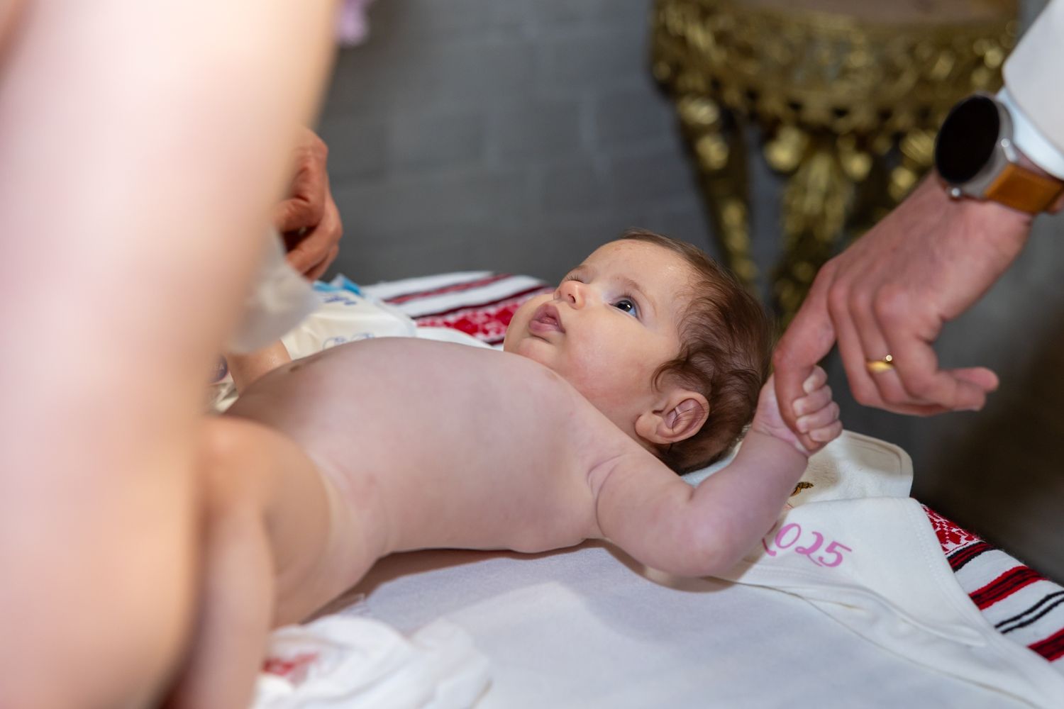 A newborn baby lying on a white cloth during a baptism ceremony, looking up curiously.
