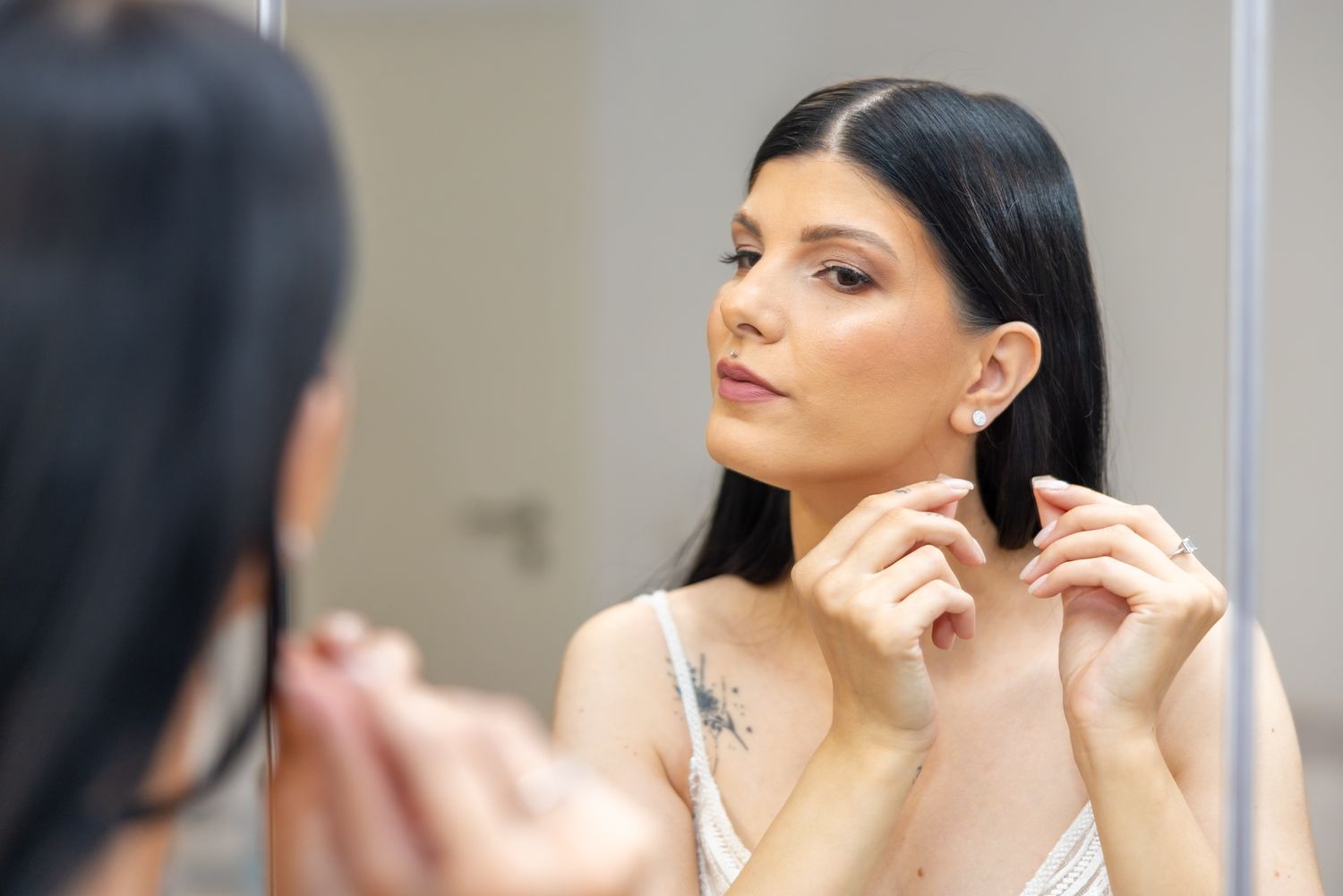 Woman putting on earrings while looking in bathroom mirror, with dark hair and tattoo visible.