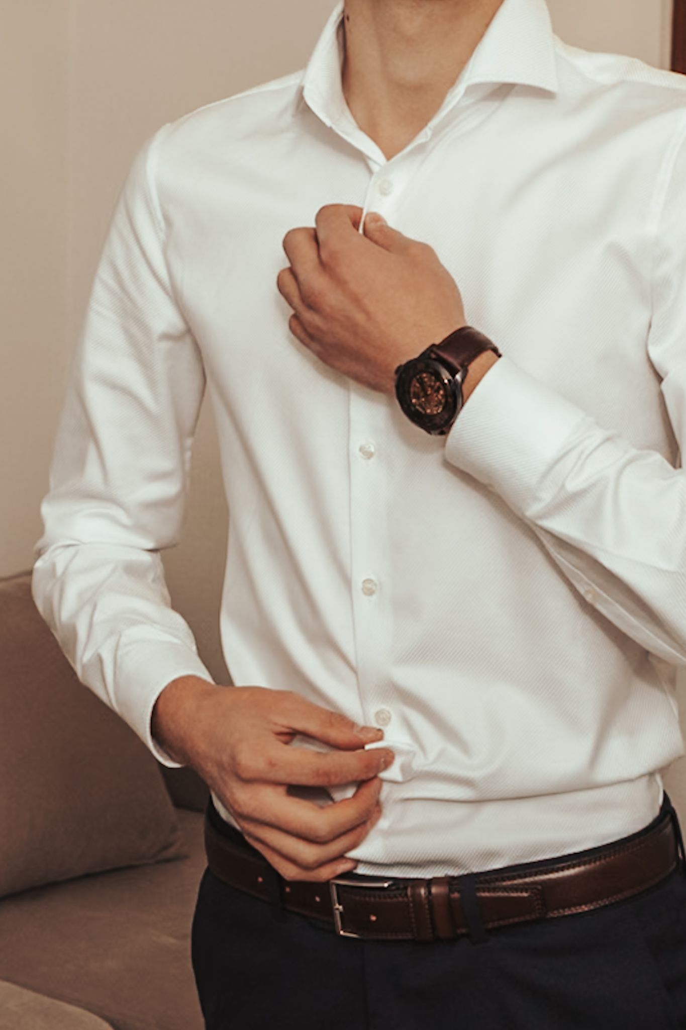 Man in white dress shirt buttoning up, wearing a brown leather watch and belt, getting dressed for formal occasion.
