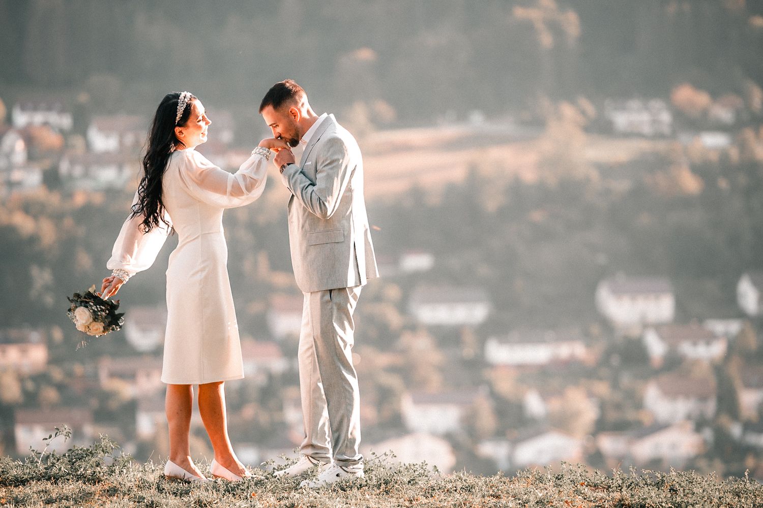 Groom kissing bride's hand on a hilltop with a soft bokeh village backdrop on their wedding day.