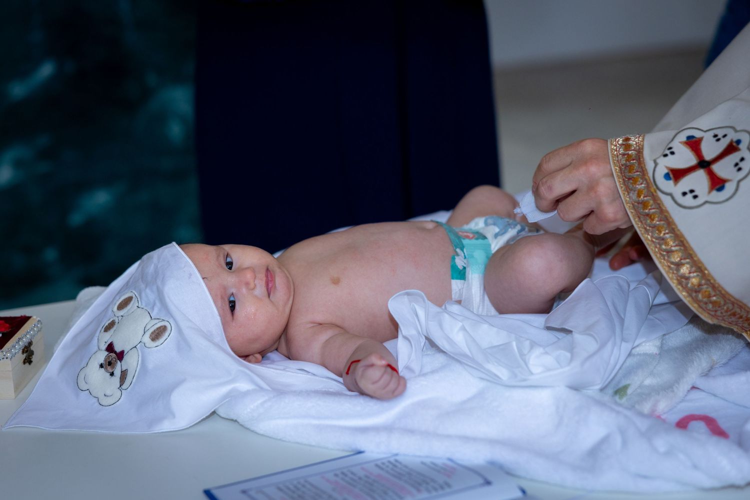 Newborn baby in white baptism outfit lying on table during Orthodox Christian christening ceremony.