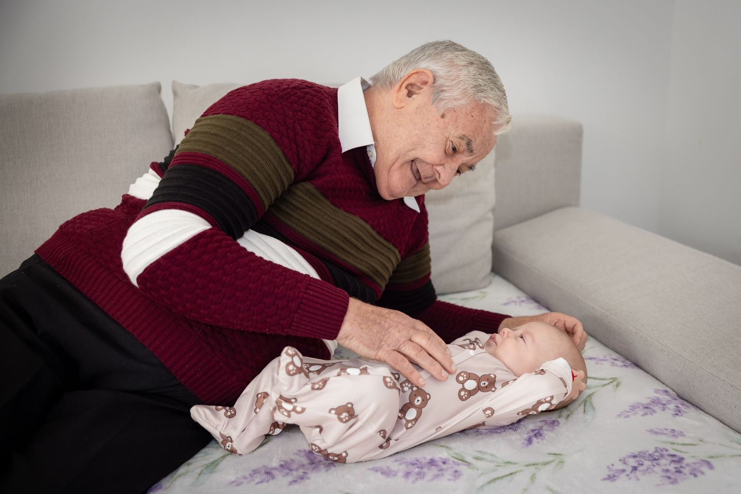 Elderly grandfather smiling and bonding with newborn baby lying on floral blanket on sofa.
