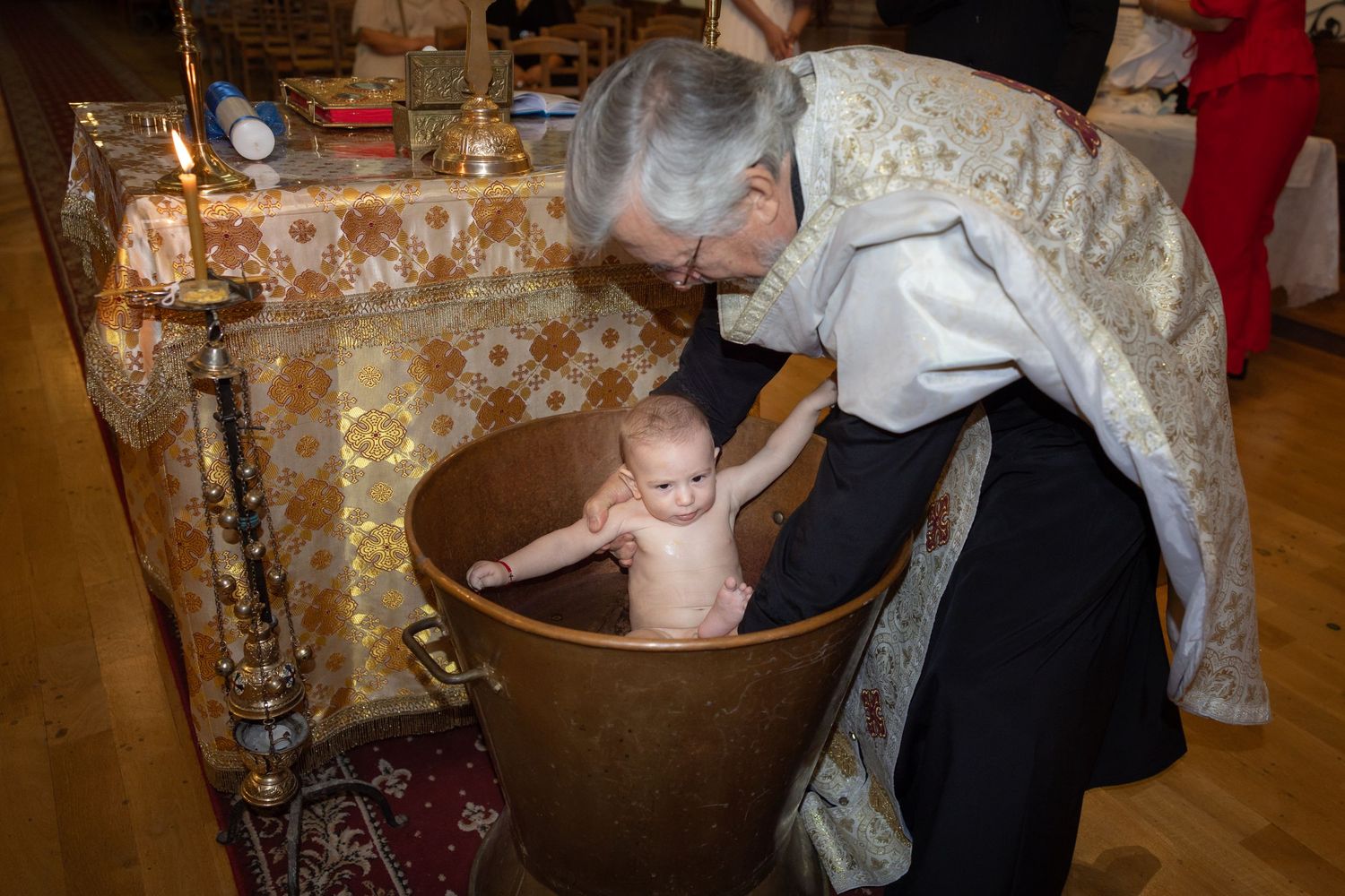 Orthodox priest performing a traditional baby baptism ceremony in a bronze font inside a church.