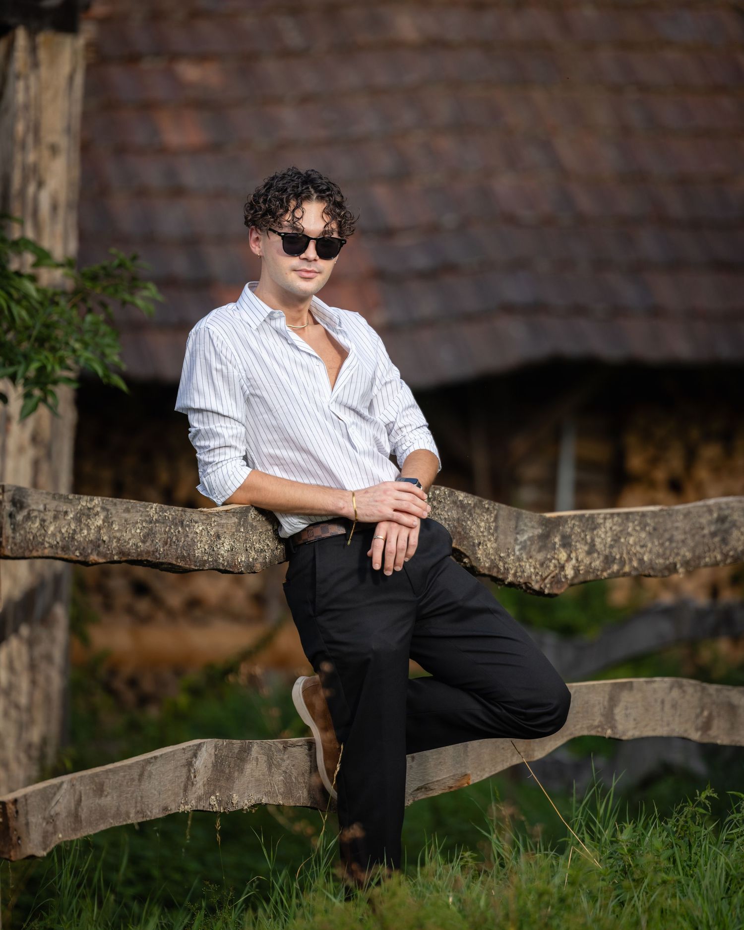 Young man in white striped shirt and sunglasses leaning on rustic wooden fence near vintage barn.