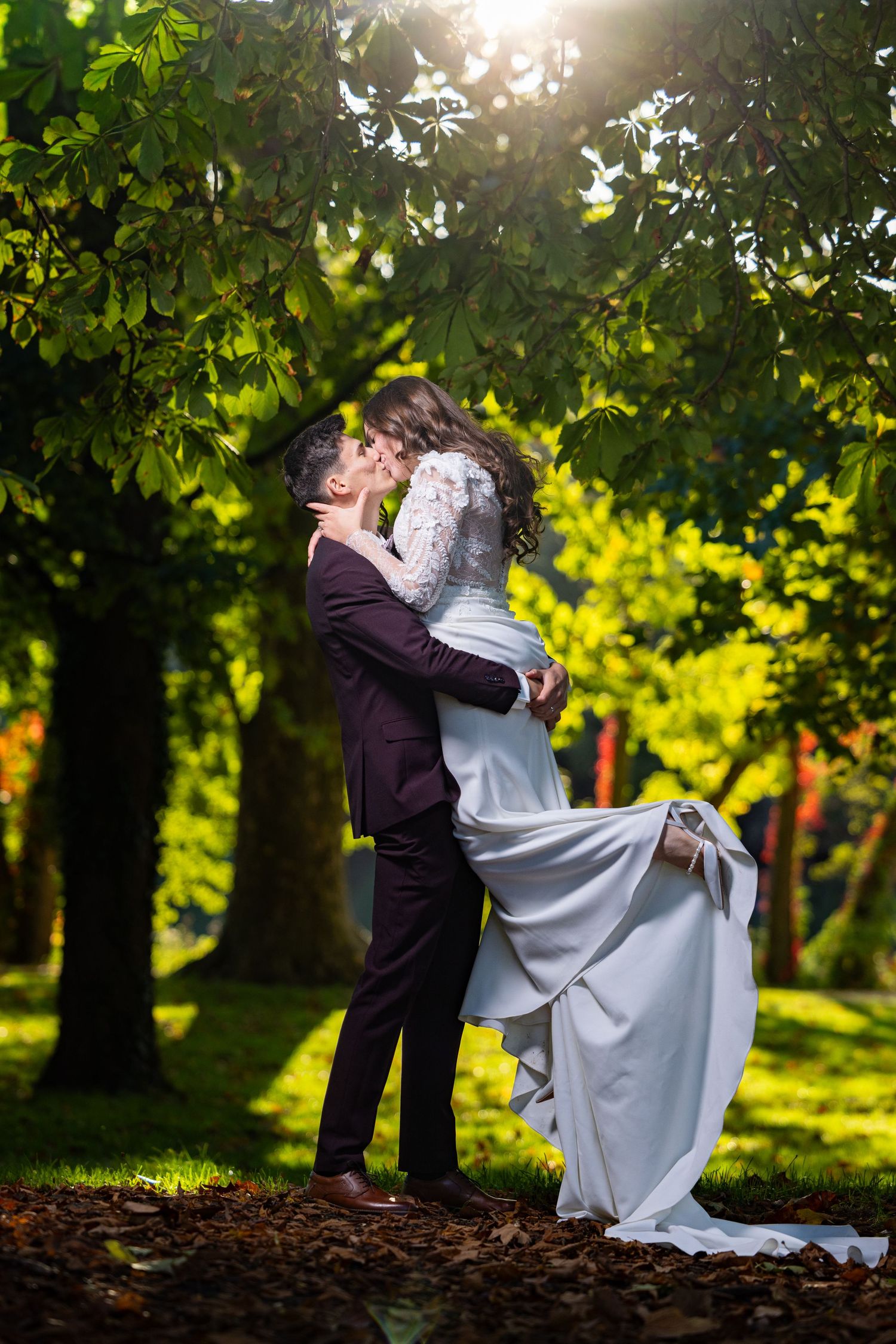 Daly hebt Emma im weißen Brautkleid hoch und küsst sie im sonnigen Park.