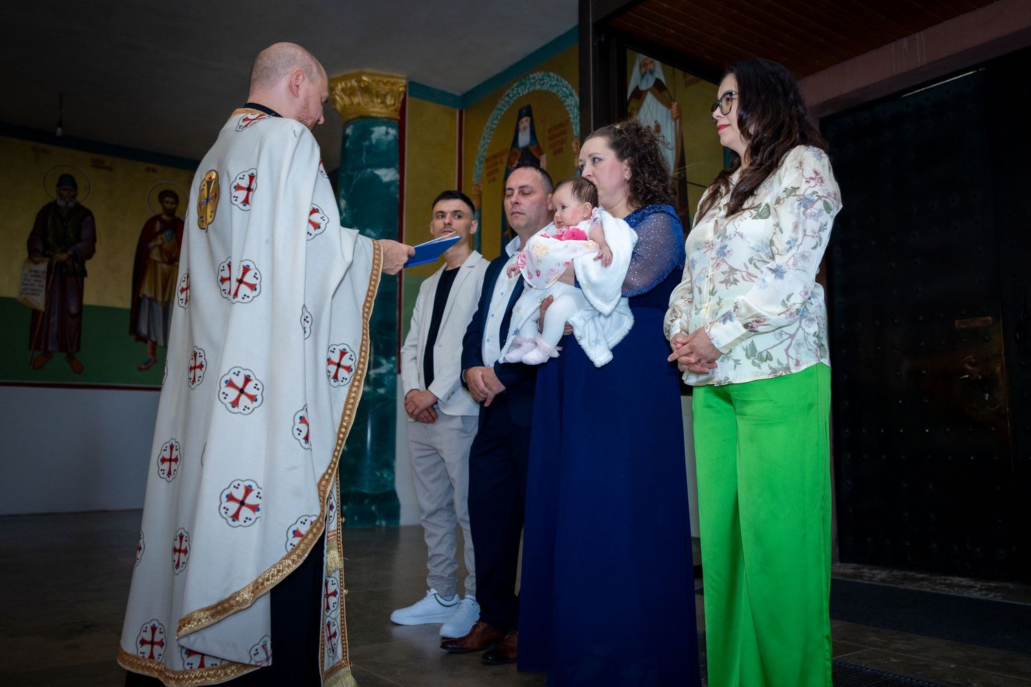 Orthodox priest performing a baptism ceremony with family members holding a baby in a decorated church.