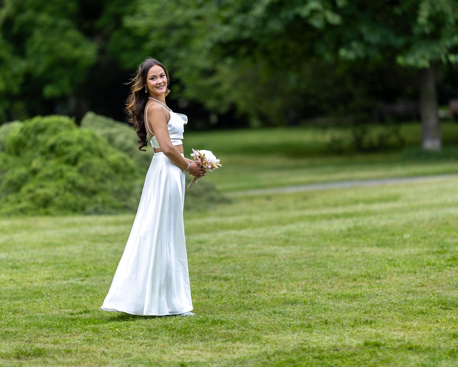 Bride in white wedding dress holding bouquet standing on green lawn outdoors.
