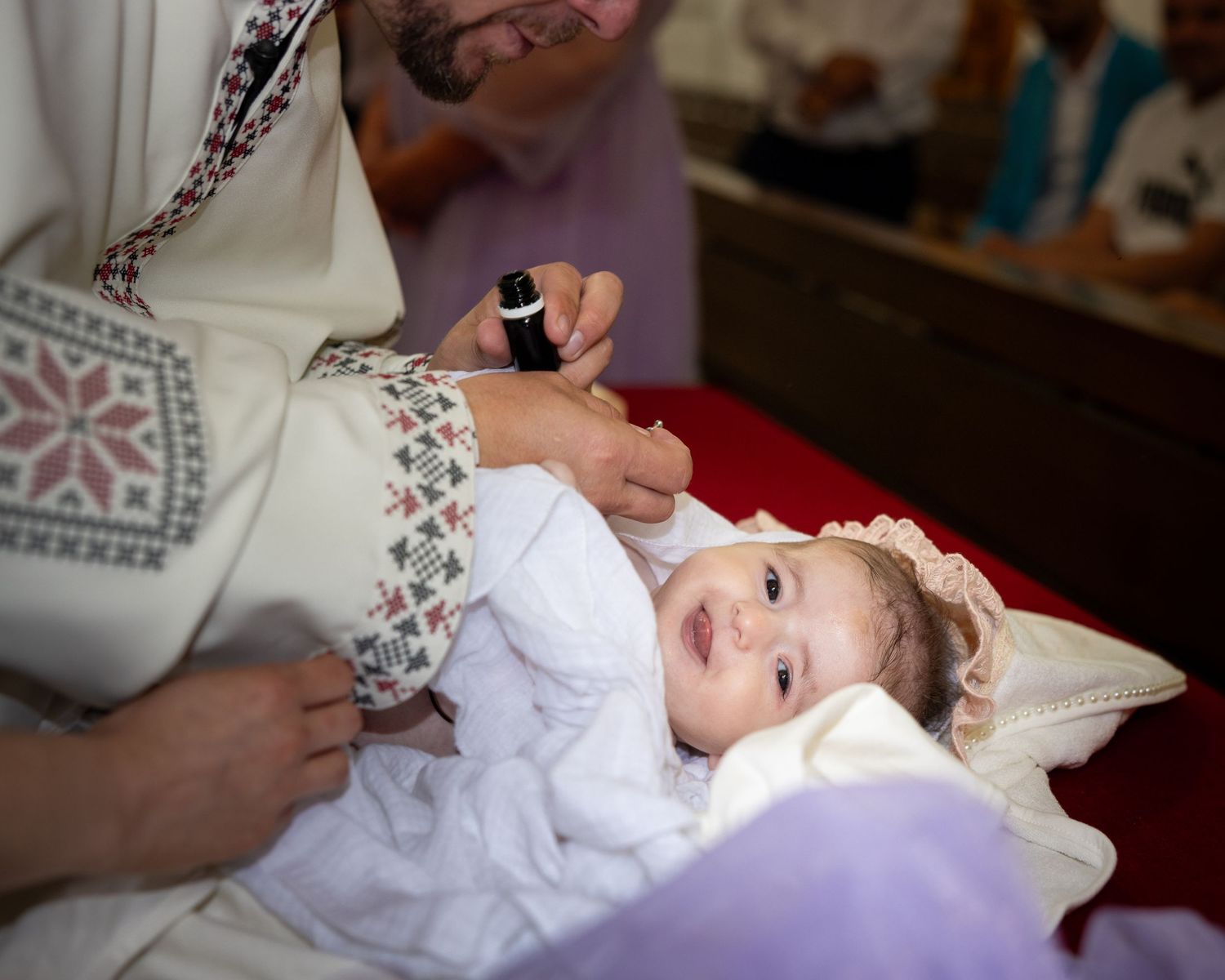 Alexia Ioana blickt während der Taufe in die Kamera, während der Priester sie auf einer weißen Unterlage salbt.