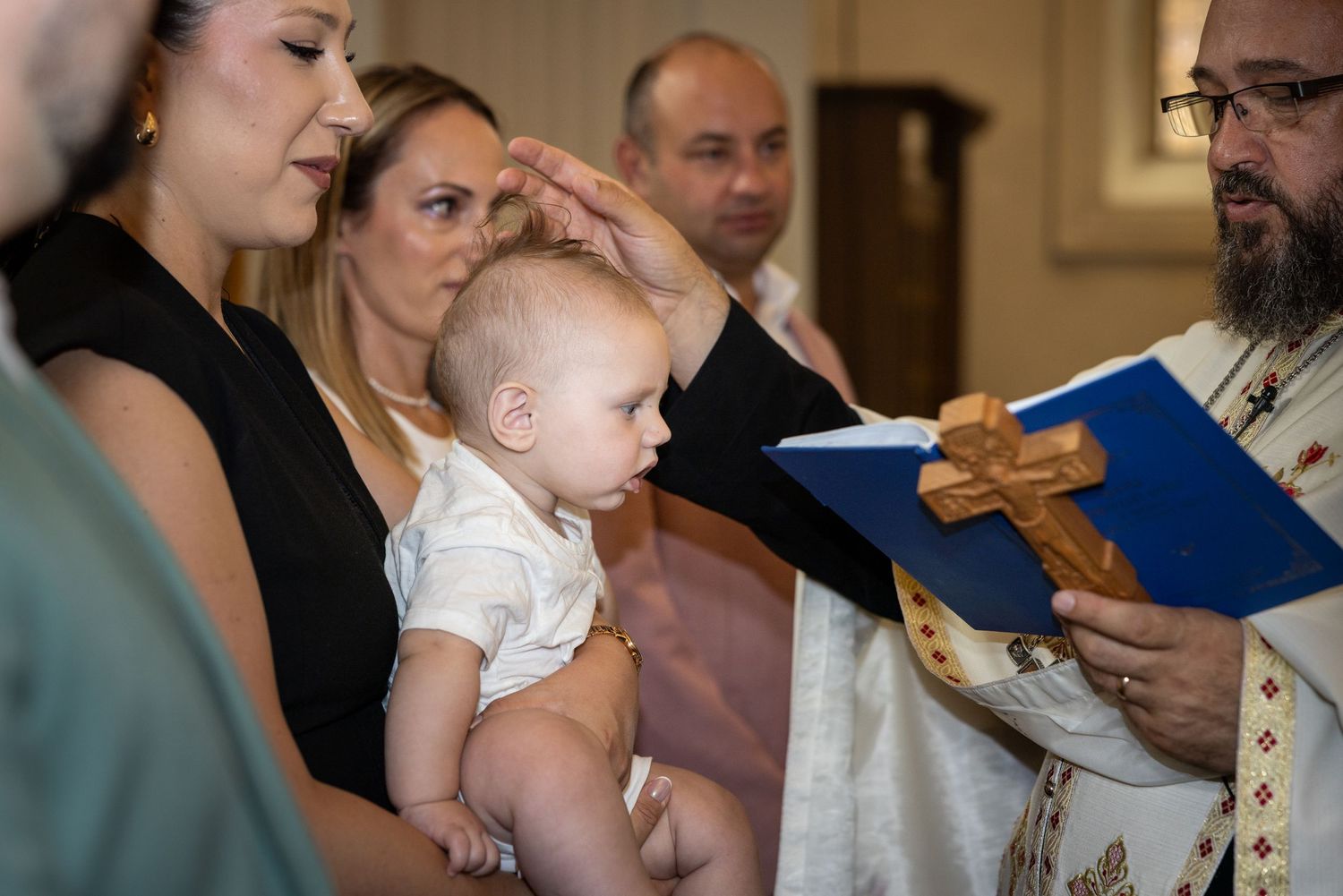 Der Priester hält die Hand über Aris’ Kopf und ein aufgeschlagenes blaues Buch mit Holzkreuz vor sich.