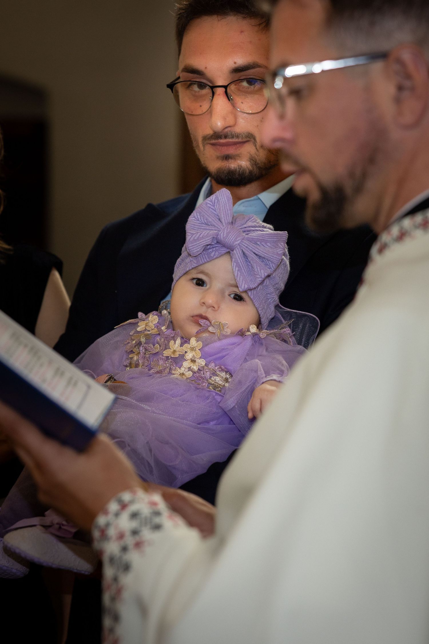 Alexia Ioana mit violetter Schleife blickt zwischen dem Mann, der sie hält, und dem Priester hervor.