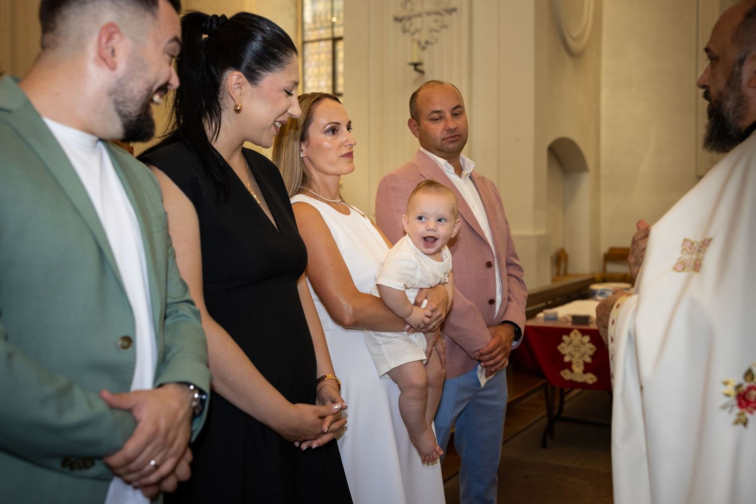 Aris lächelt auf dem Arm einer Frau im weißen Kleid, umgeben von den Erwachsenen bei der Taufe.