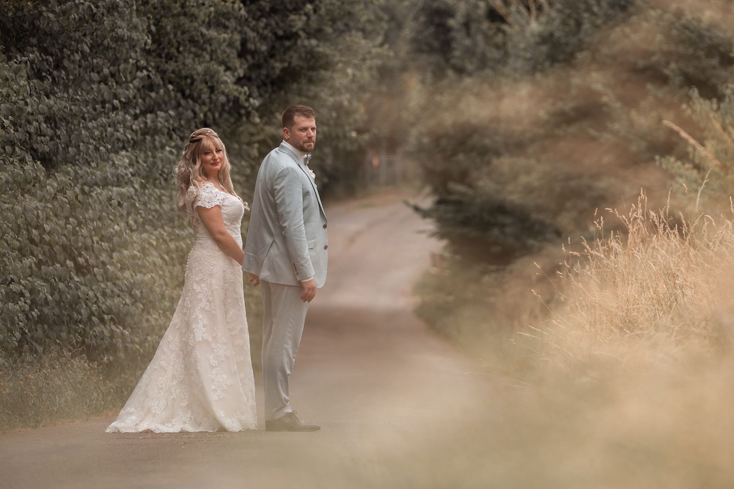 Bride in lace gown and groom in grey suit holding hands on a rustic country road.