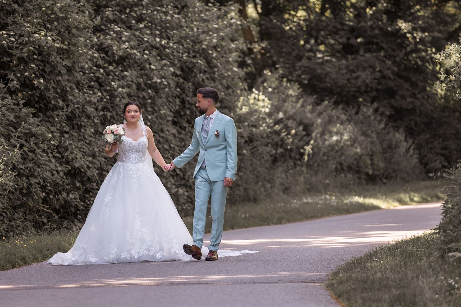 Bride and groom walk hand-in-hand on a scenic tree-lined path, bride in white gown, groom in light blue suit.