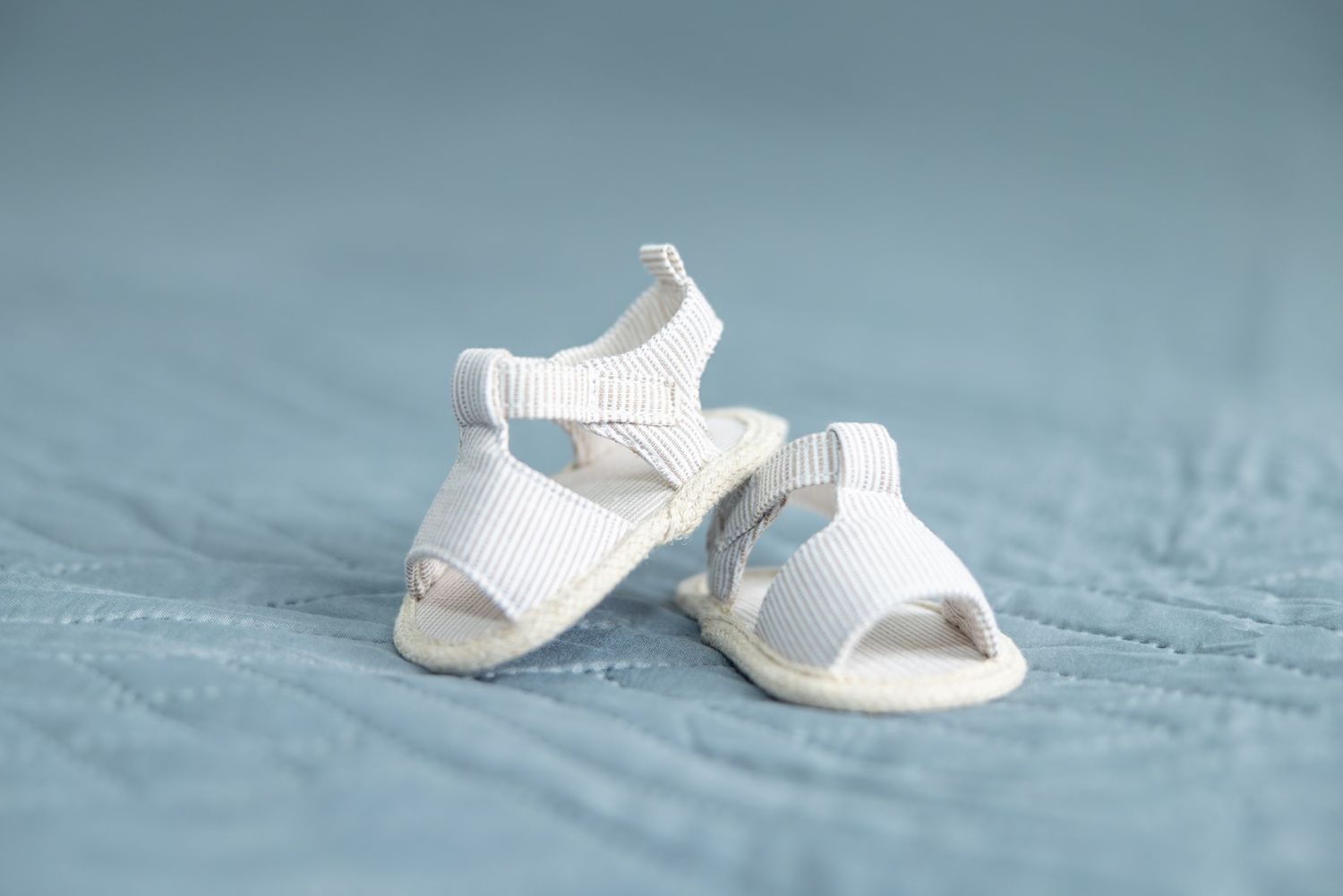 Tiny white baby sandals with ribbed straps displayed on a soft blue quilted background.