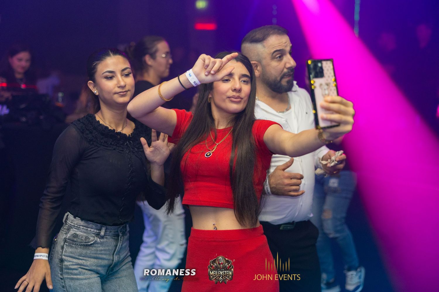 Eine Frau im roten Oberteil hält ein Smartphone für ein Gruppenfoto im violett beleuchteten Club Meilenstein.