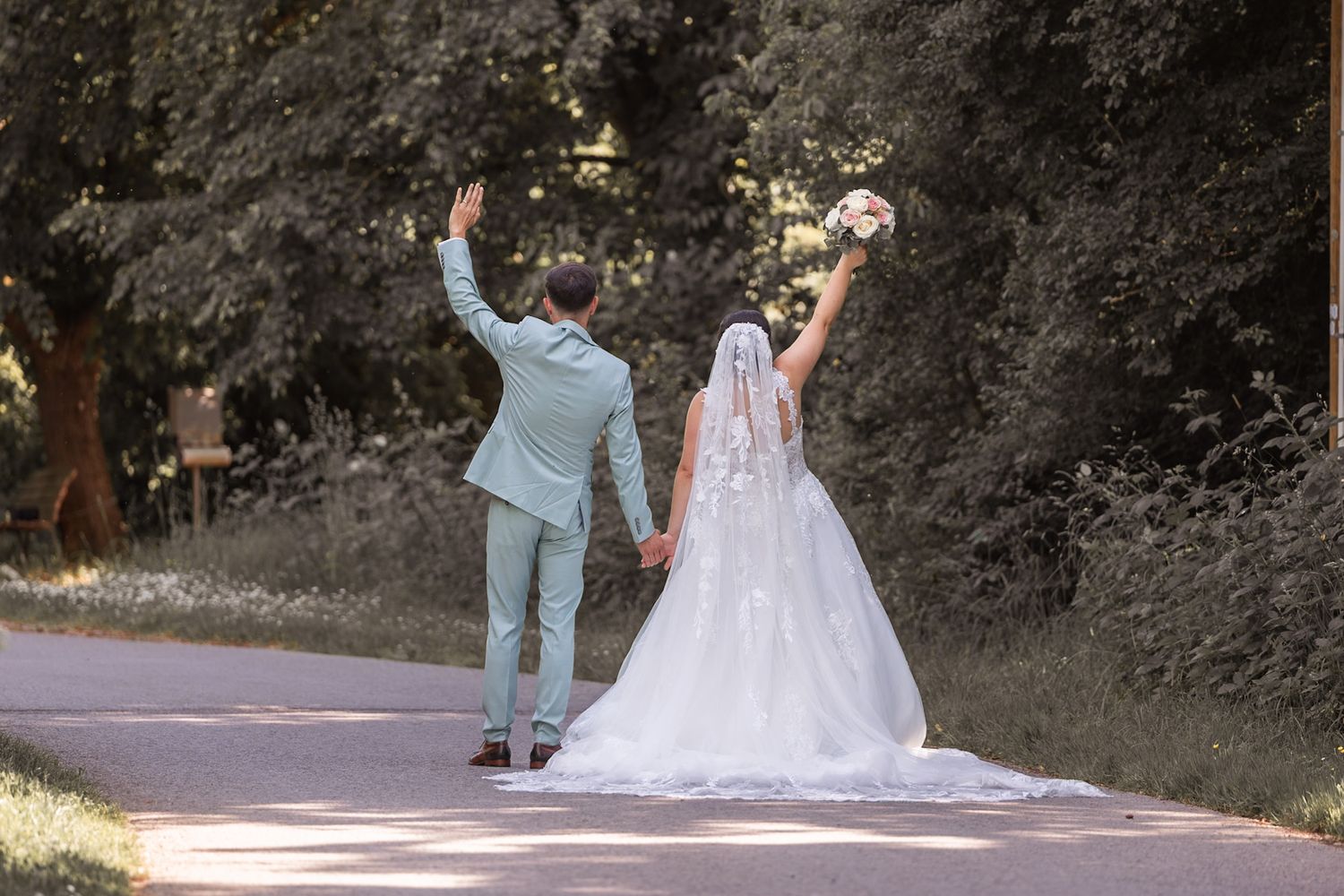 Bride and groom walking hand in hand, raising arms joyfully on a tree-lined path in wedding attire.