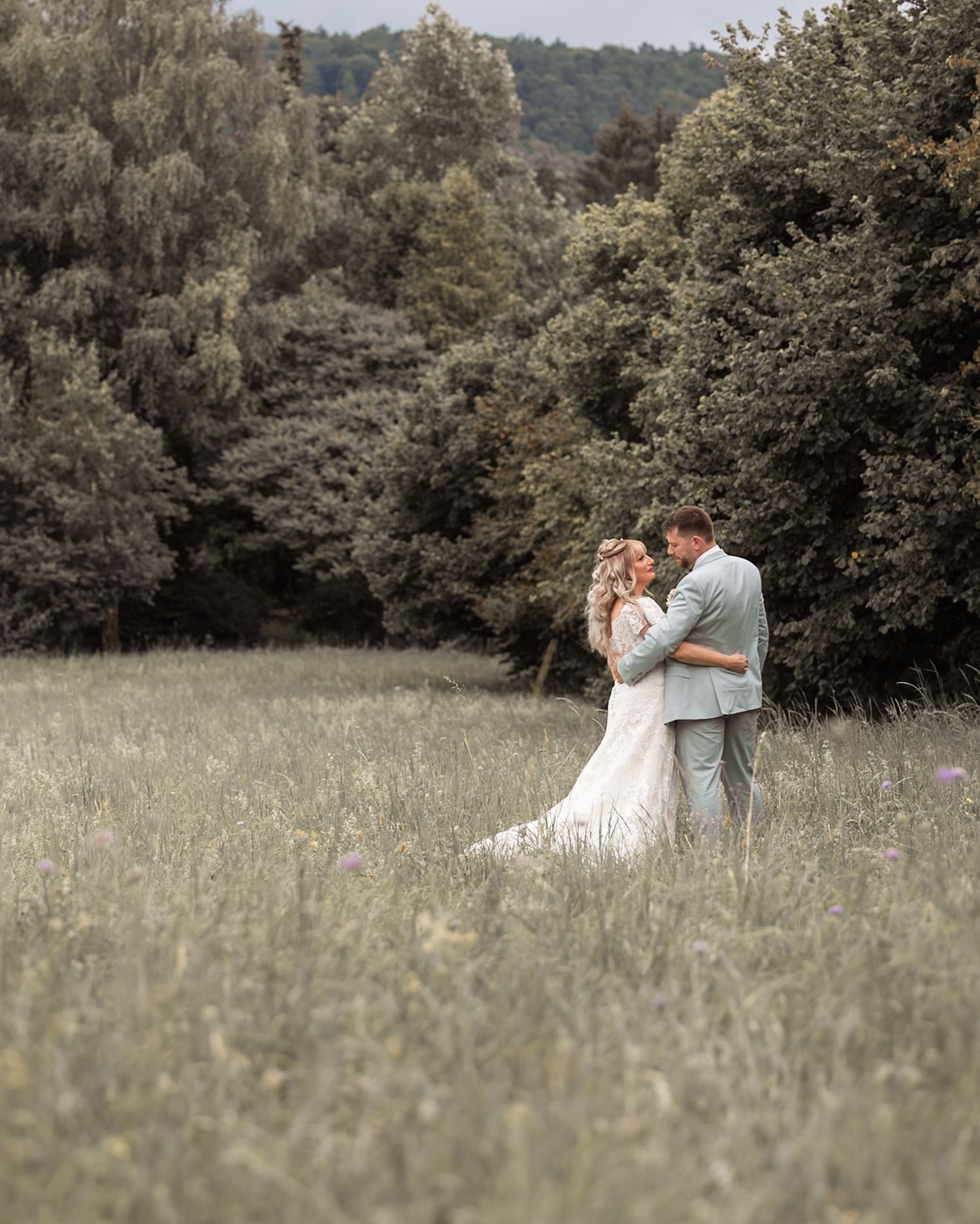 Newlywed couple embracing in a sunlit meadow surrounded by trees, bride wearing a flowing white dress.