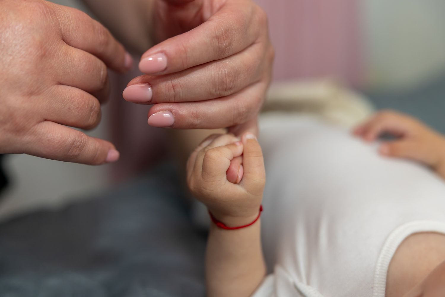 A newborn baby grasping an adult finger, wearing a red bracelet, while lying on a white surface.
