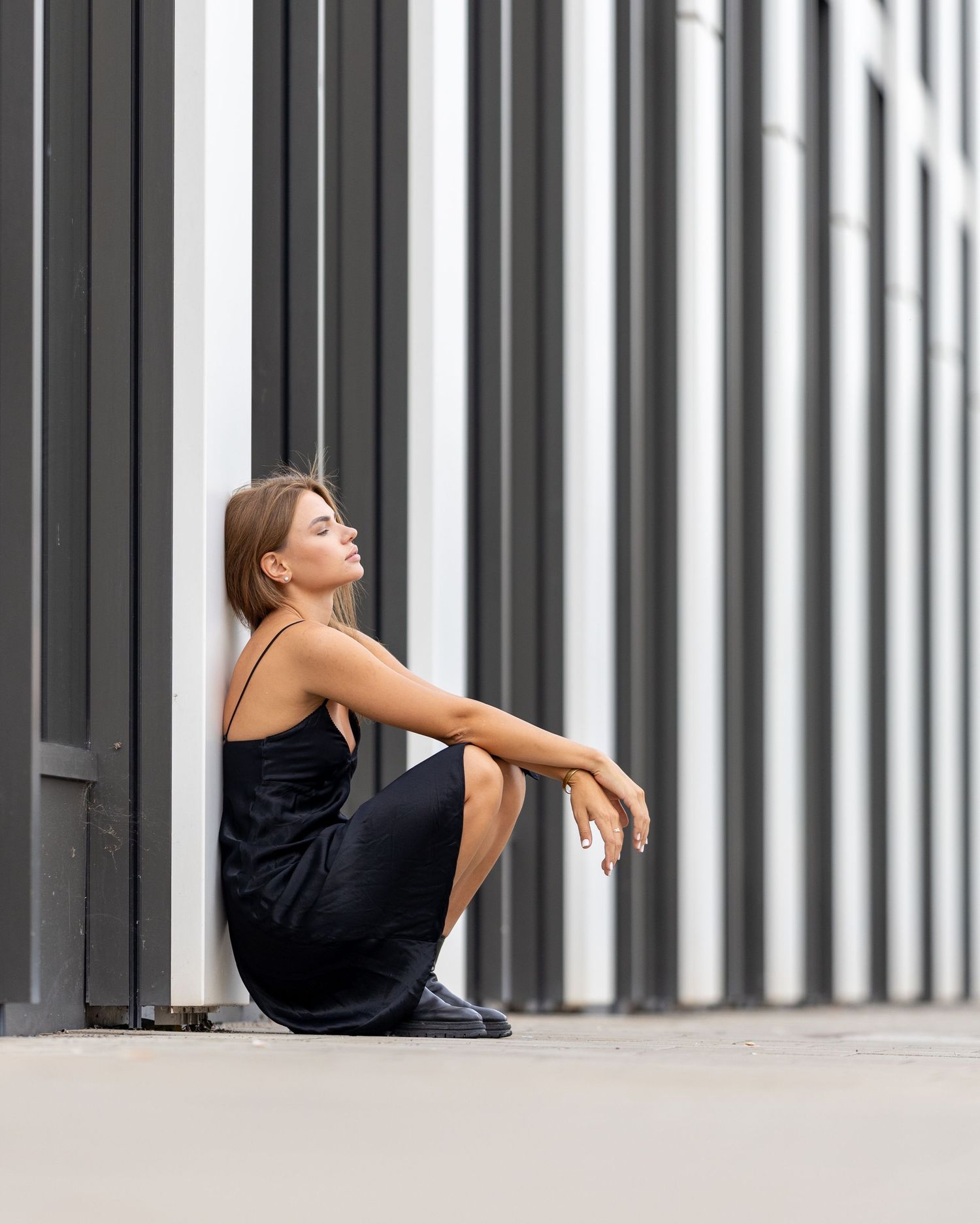 Woman in black dress sitting against striped black and white wall outdoors, looking up thoughtfully.