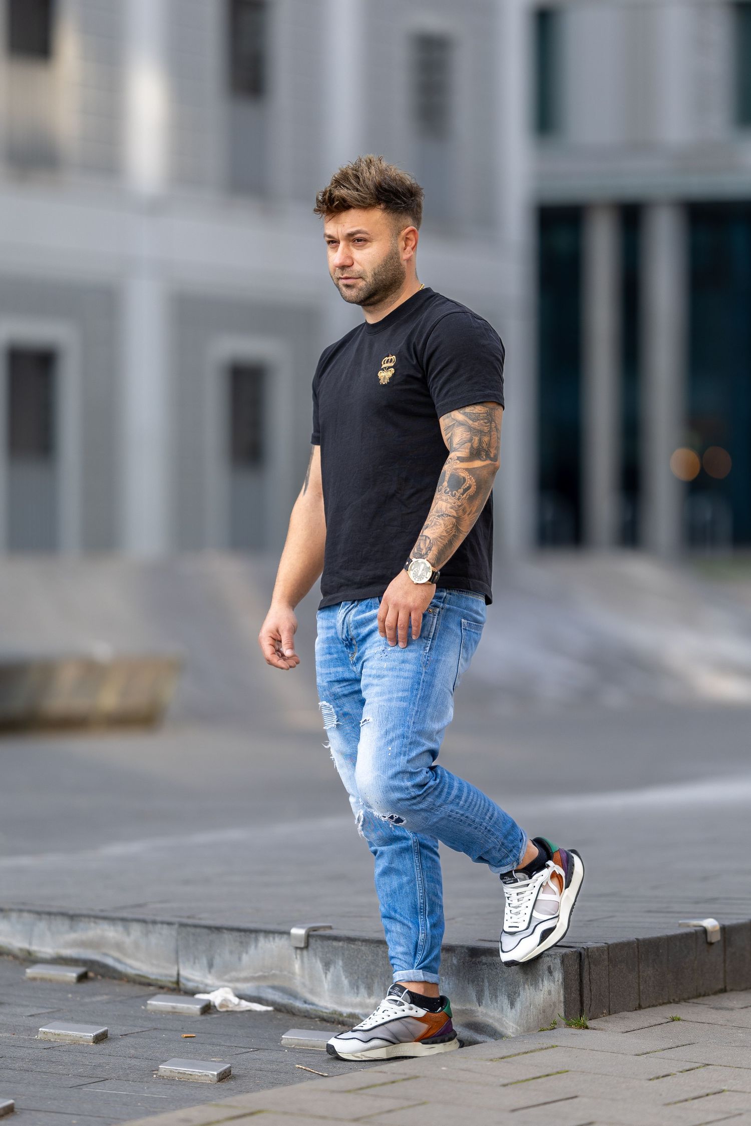 Tattooed man in black t-shirt, blue jeans, and sneakers walking outdoors near a modern building.