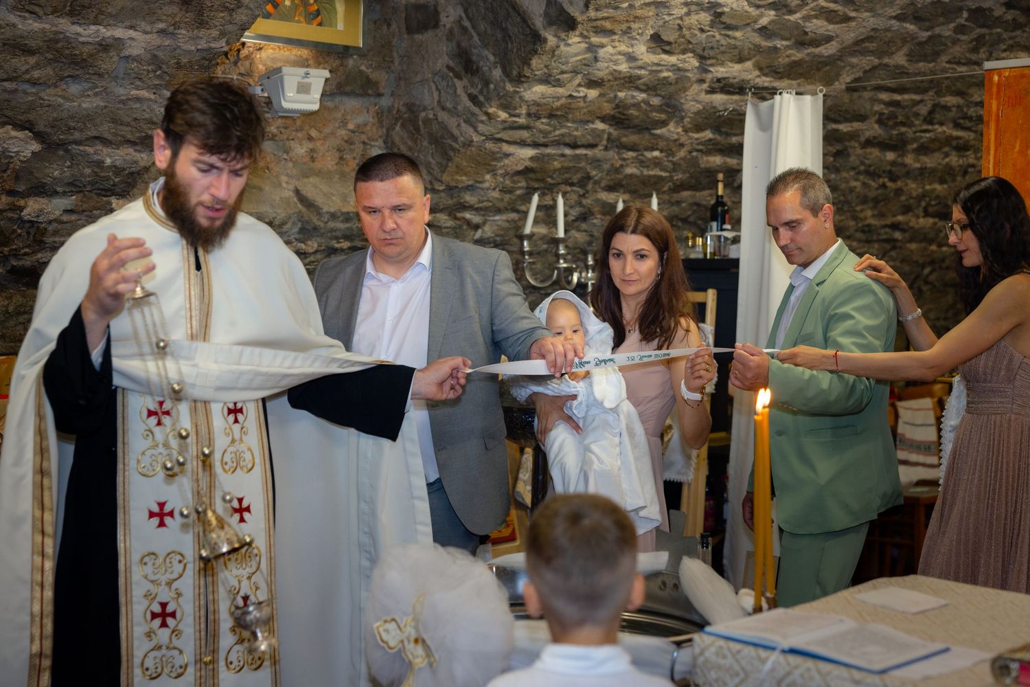 Der Priester steht mit Milan, mehreren Begleitpersonen und einem Kind am Taufbecken.