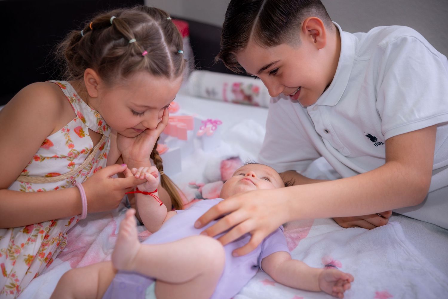 Two older siblings lovingly interact with a newborn baby lying on a bed, sharing a tender family moment.