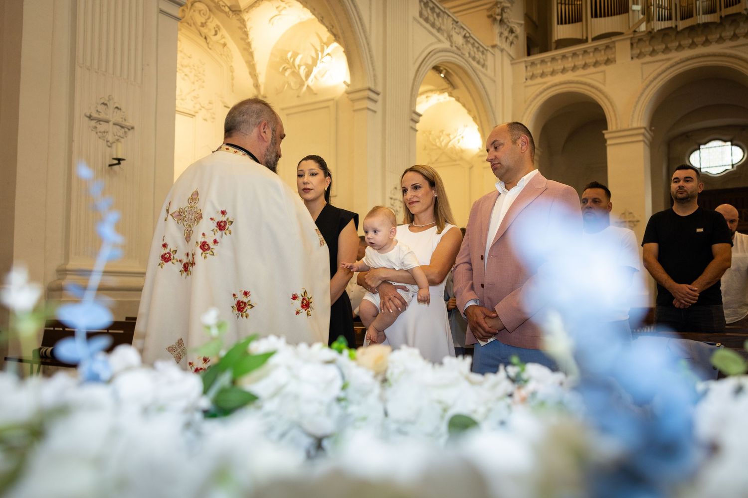 Aris wird vor dem Priester auf dem Arm gehalten; weiße und blaue Blumen rahmen die Szene im Vordergrund.