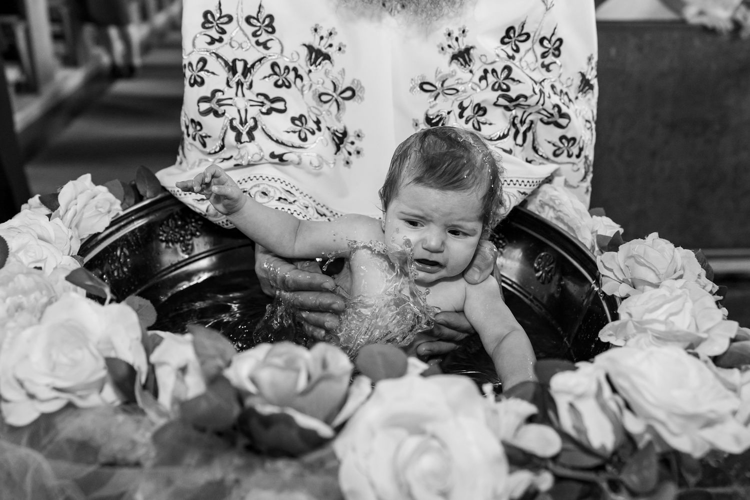 Black and white photo of a baby being baptized in a font surrounded by white roses during a church ceremony.
