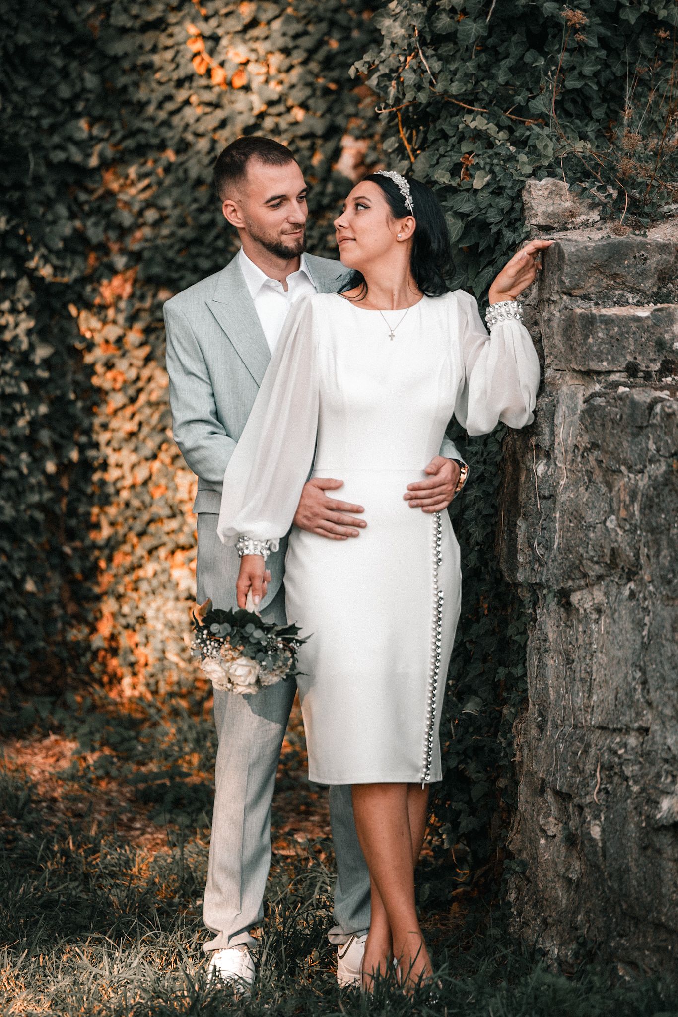 Elegant couple in white formal attire embracing by ivy-covered stone wall during outdoor wedding photoshoot.