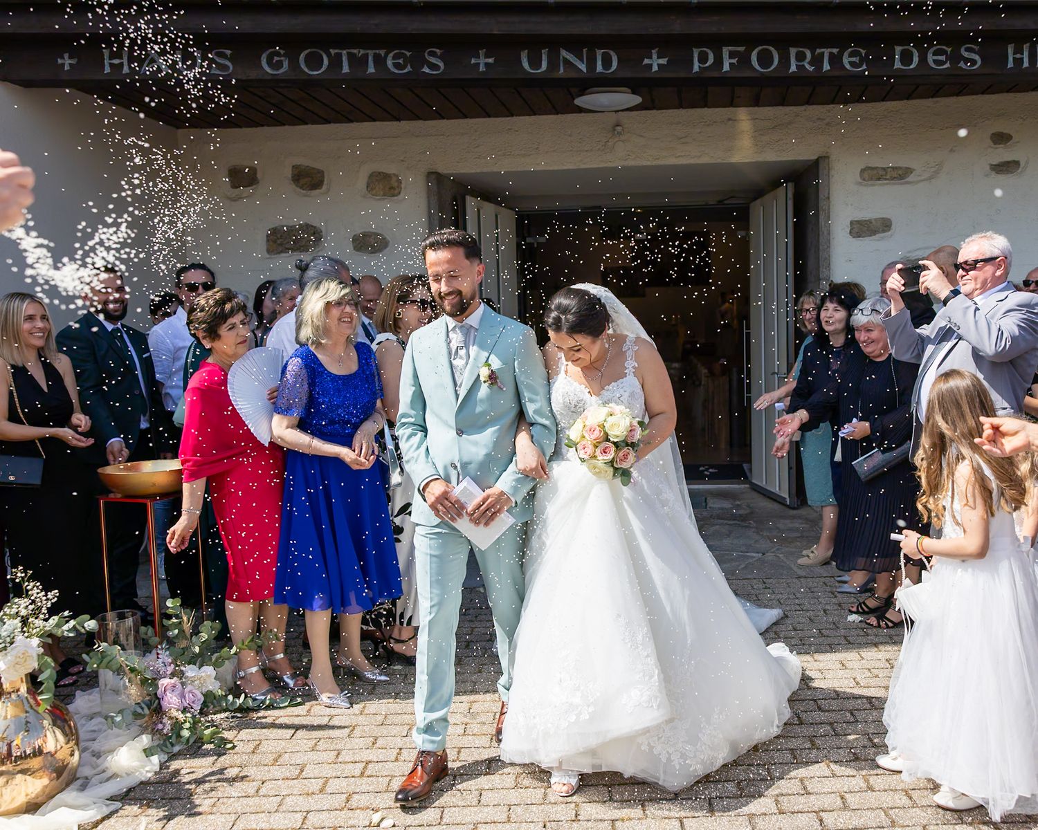 Joyful newlyweds exit a church as guests throw confetti, bride in white gown holding bouquet, groom in light blue suit.