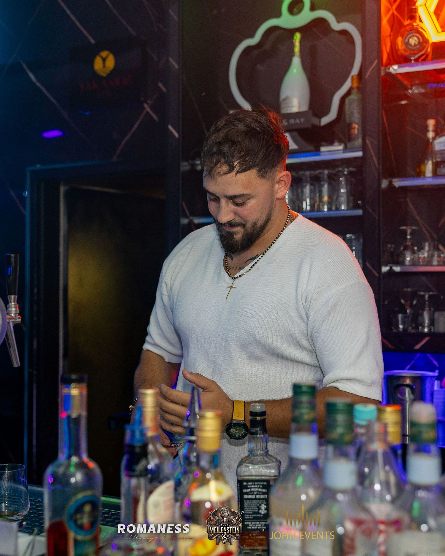 Ein Mann im weißen T-Shirt steht hinter einer Reihe von Flaschen an der Bar des Clubs Meilenstein.