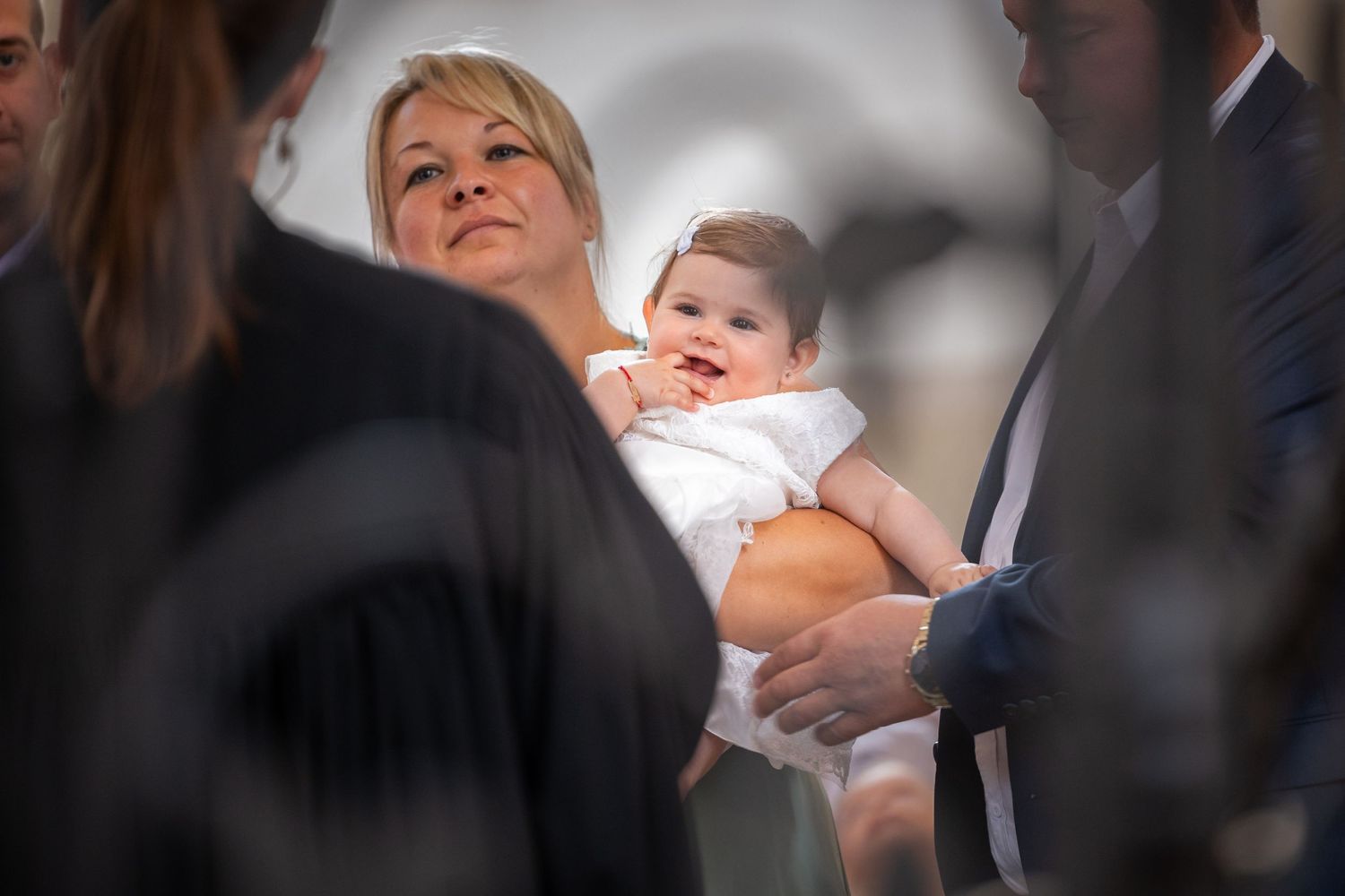 Karoline lächelt im weißen Taufkleid auf dem Arm ihrer Patin während der Zeremonie in der Kirche.