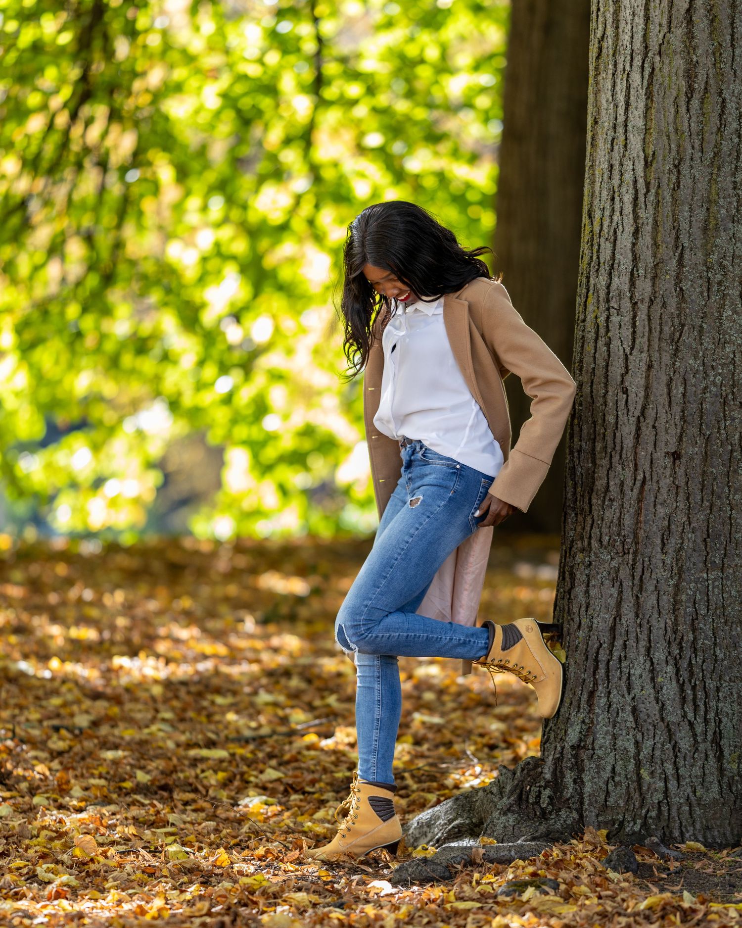 Jacky lehnt in hellem Mantel an einem großen Baum im Park