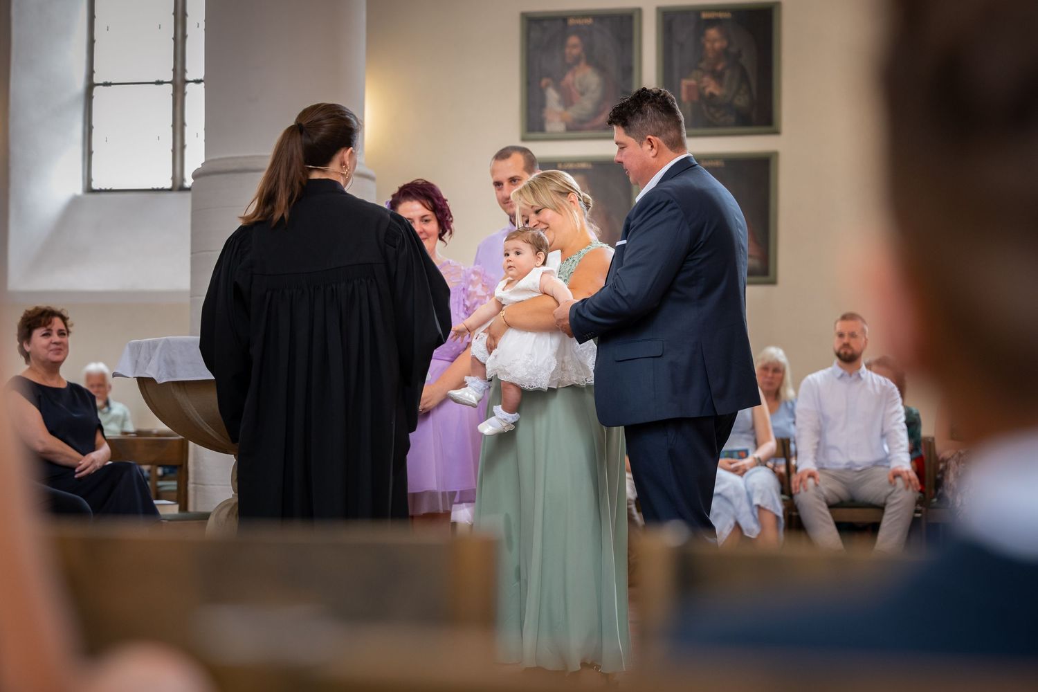 Karoline mit ihren Eltern und Paten während der Taufe, durch die Kirchenbänke fotografiert.