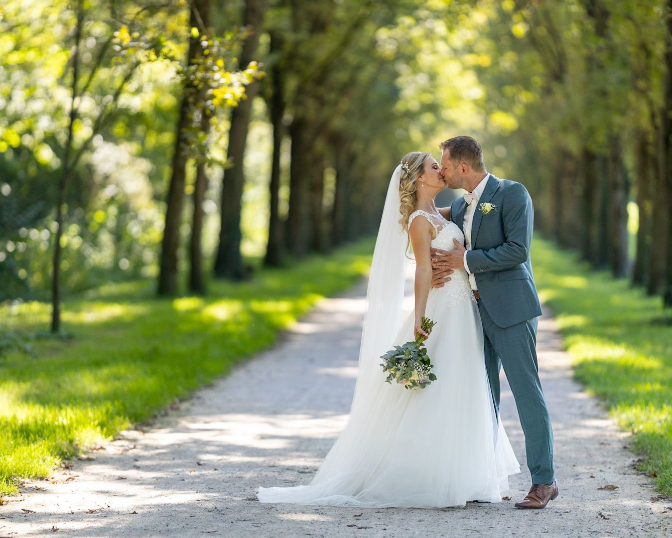 Bride and groom sharing a romantic kiss on a tree-lined path, bride holding a bouquet in a white gown.