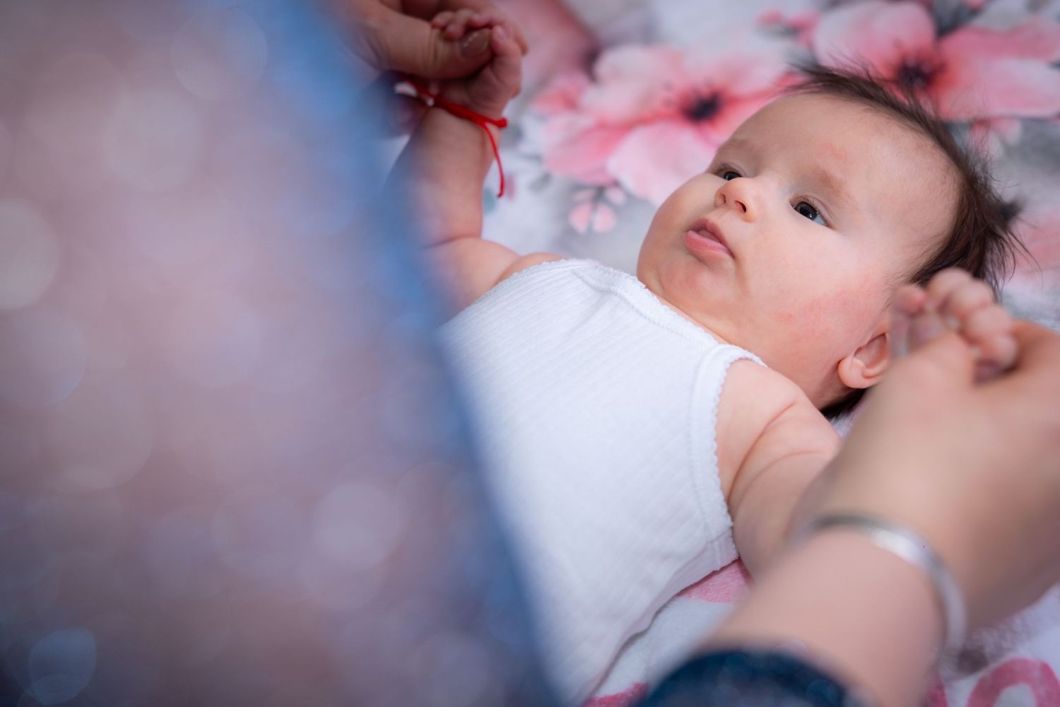 Newborn baby lying on floral blanket in white onesie, gazing upward while adult hands gently hold a red string nearby.