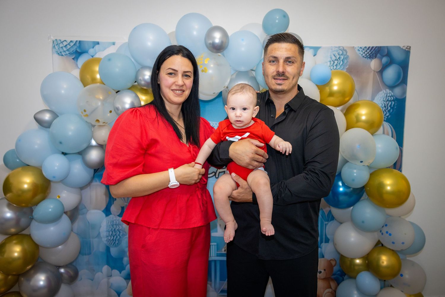 Carlo im Arm eines Mannes neben einer Frau in Rot, vor blau-goldener Ballondekoration bei seiner Tauffeier in Straßburg.