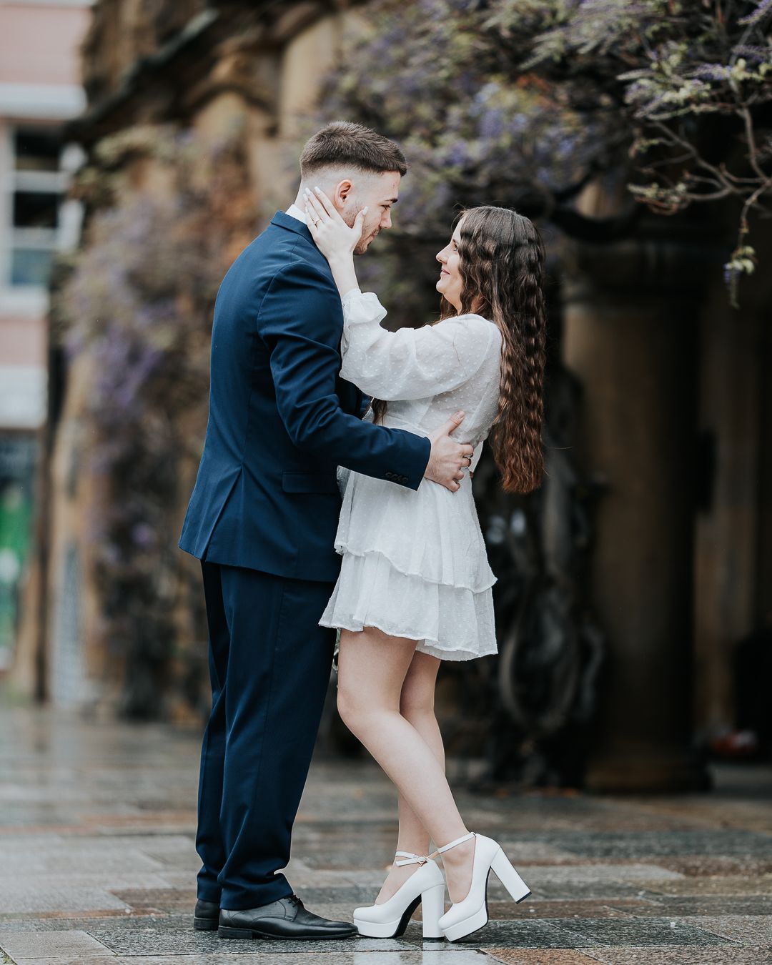Couple in love embracing outdoors, man in navy suit, woman in white mini dress and heels, with wisteria blooms behind them.