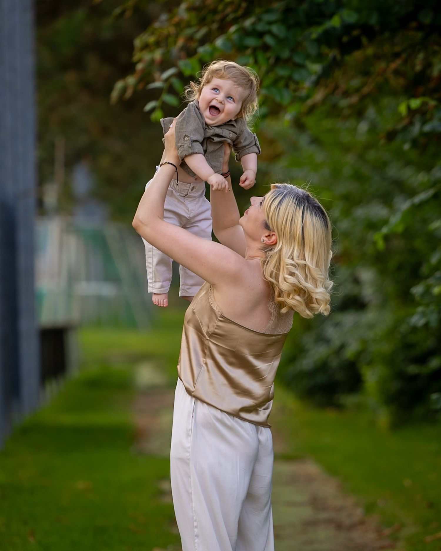 A joyful mother lifts her laughing baby overhead outdoors, both wearing neutral tones against a lush green background.