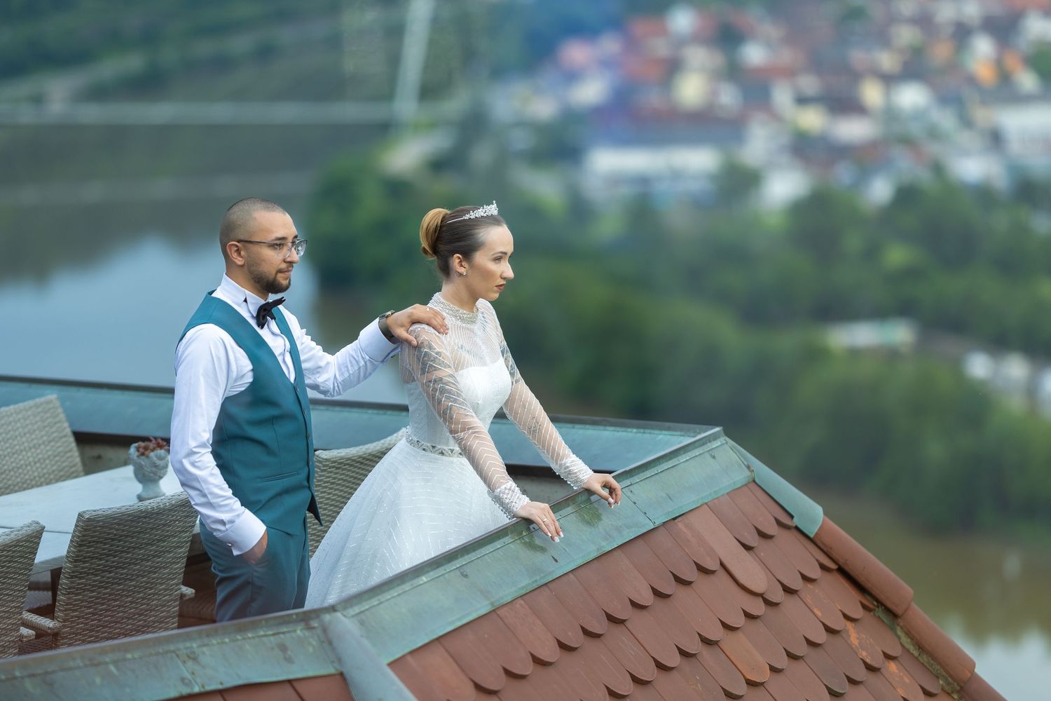 Bride and groom stand on rooftop with river view, groom in teal vest touches bride's shoulder in white lace gown.