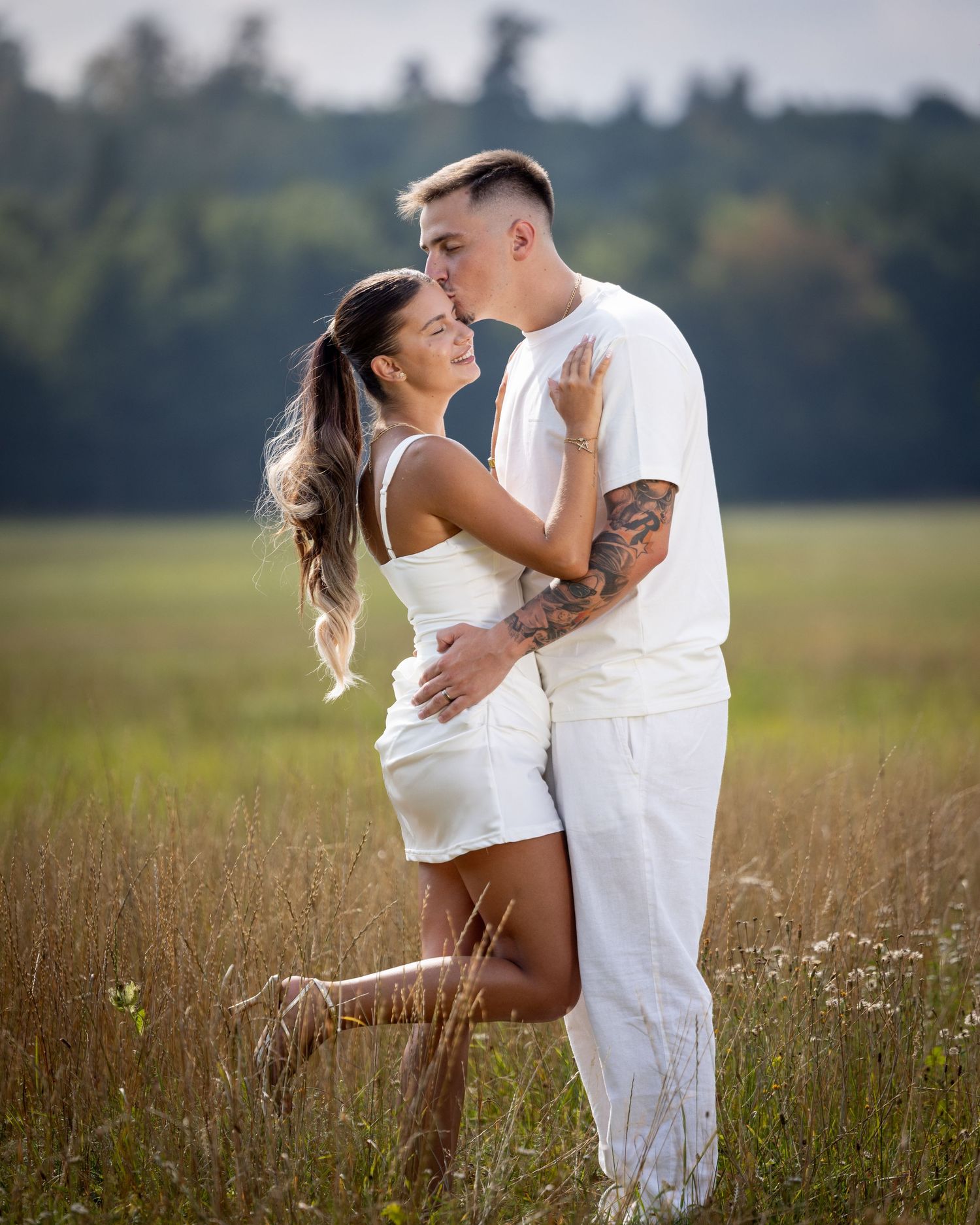 Lorena und Bogdan stehen in heller Kleidung eng beieinander auf einer Wiese und berühren sich mit den Lippen.