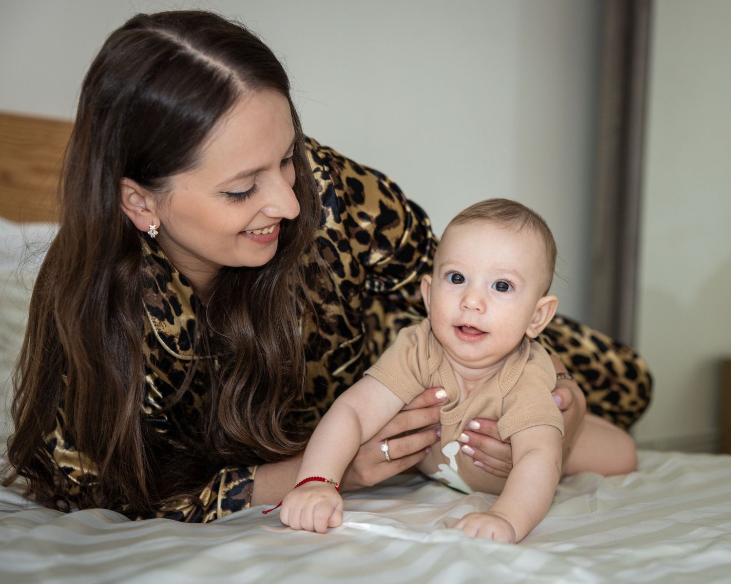 A smiling mother in leopard print top lying on a bed with her curious baby during tummy time.