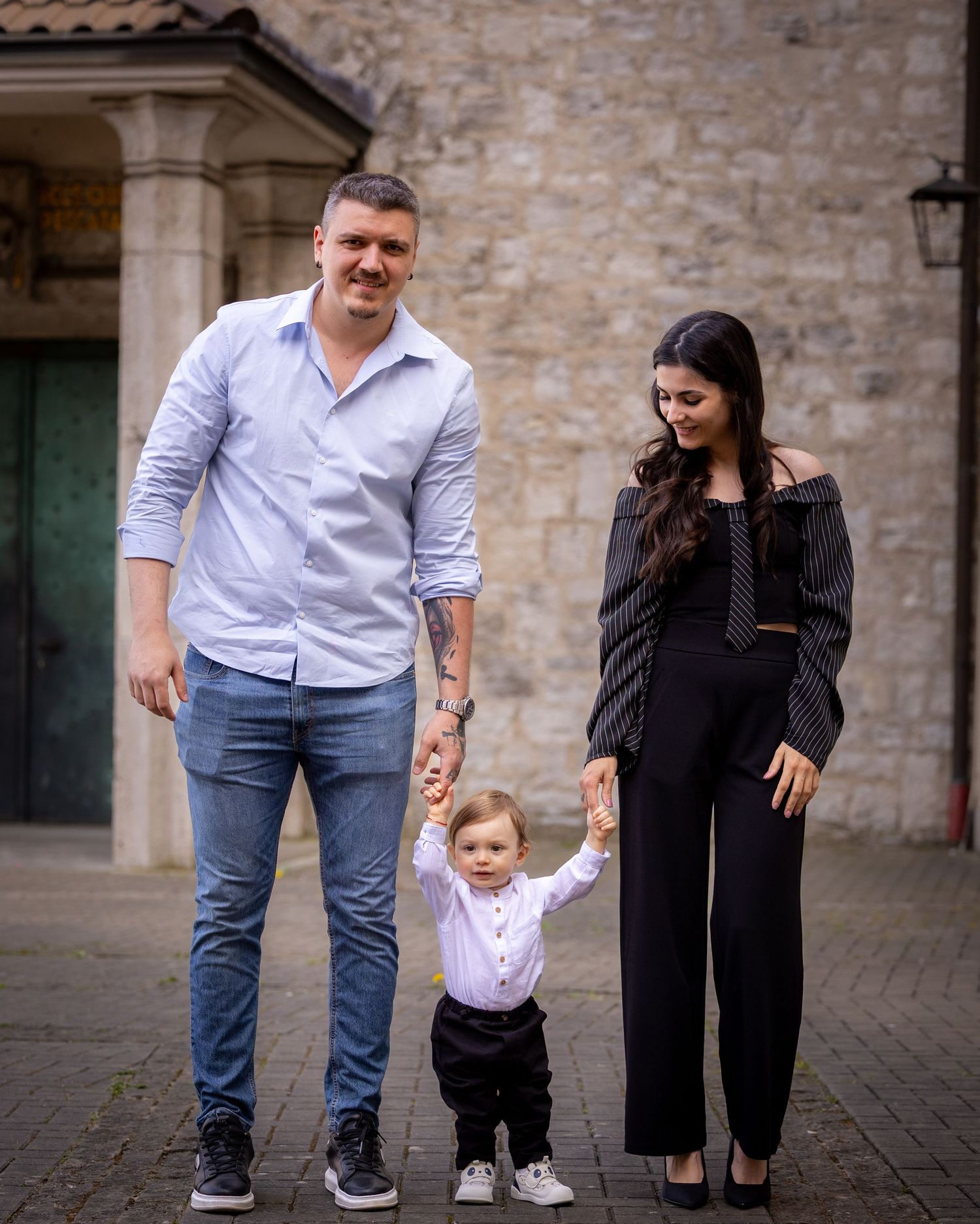 Family portrait of parents holding toddler's hands outdoors near stone building.