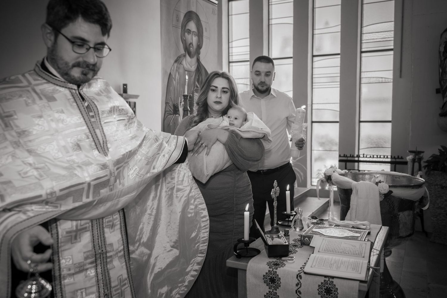 Schwarz-Weiß-Aufnahme des Priesters mit Buch und ausgestreckter Hand vor der Familie mit Lara und Taufkerze.
