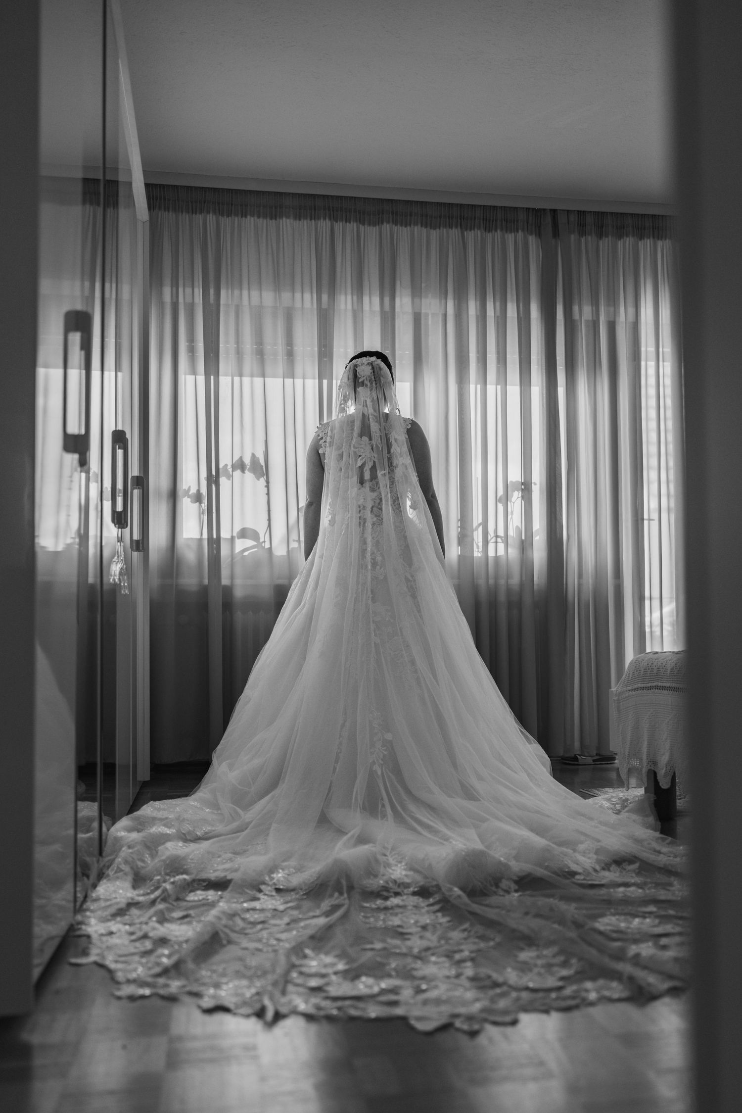 Die Braut steht im Hochzeitskleid mit langer Schleppe und Schleier vor einem hellen Fenster.