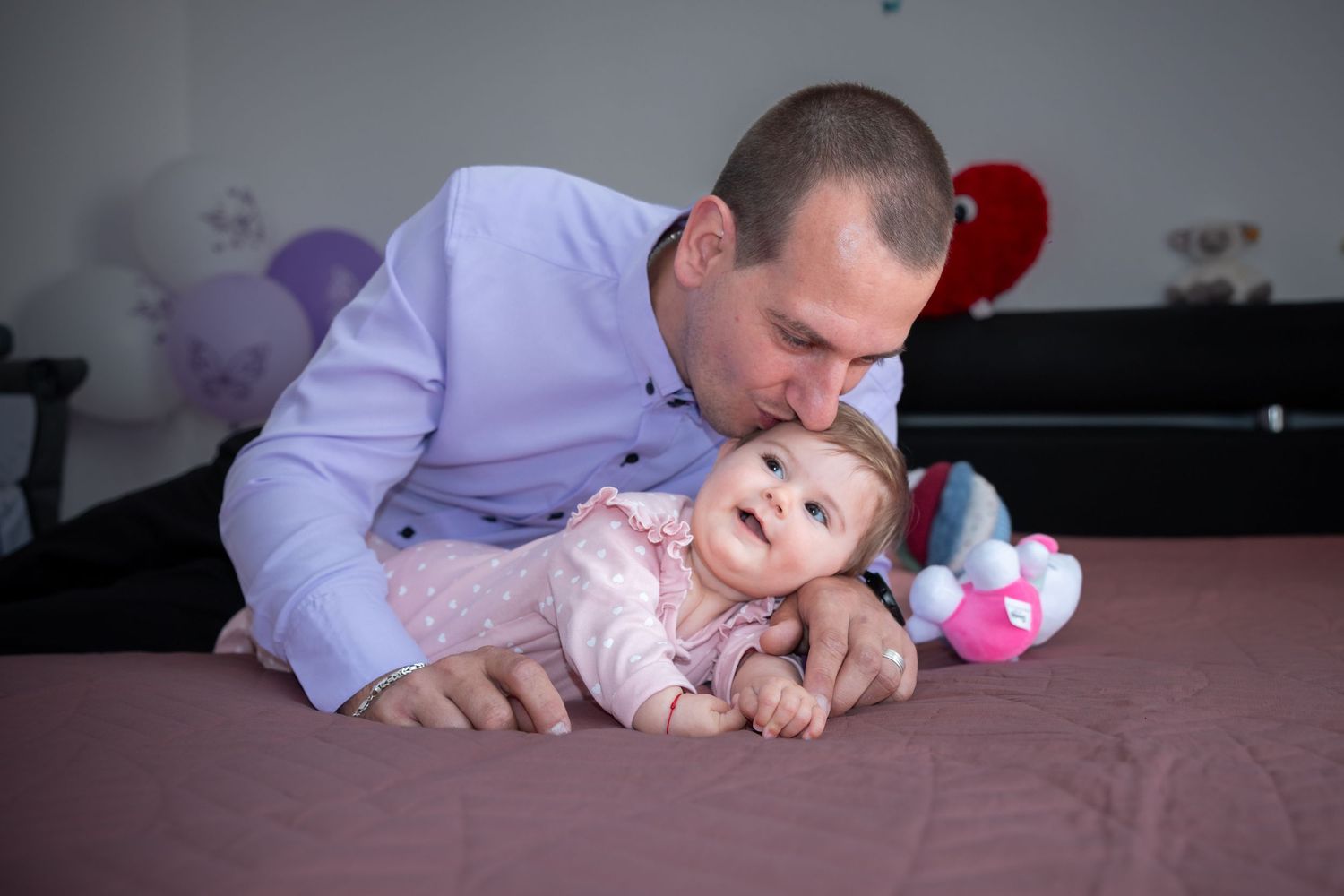 Karolines Vater küsst ihre Stirn, während sie in rosa Kleidung auf dem Bett liegt.