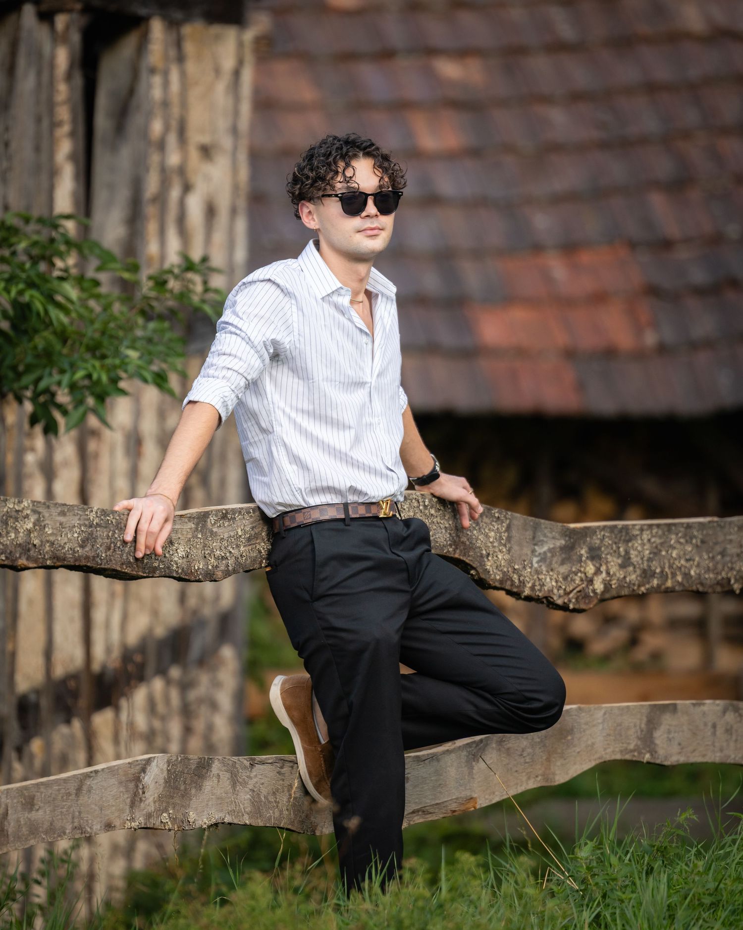 Young man with curly hair and sunglasses leaning on a rustic stone fence near a vintage wooden building.