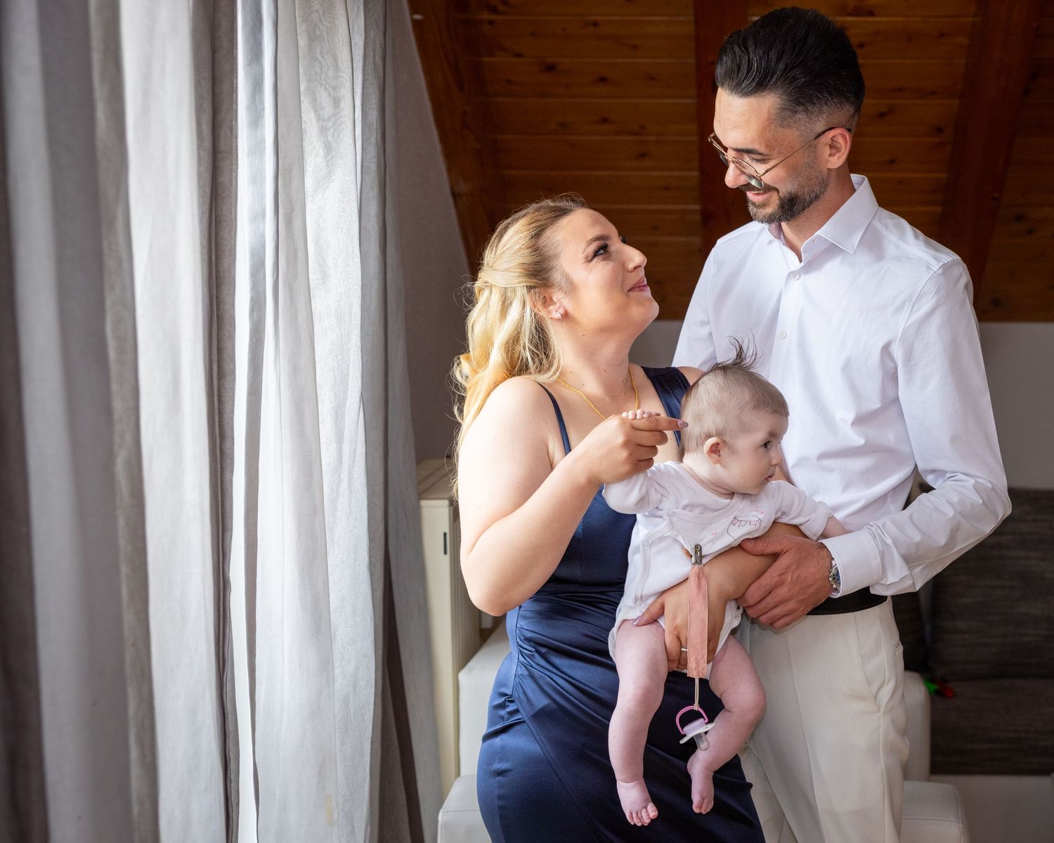 A couple in formal attire smiles warmly while holding their baby near a staircase in a bright indoor setting.